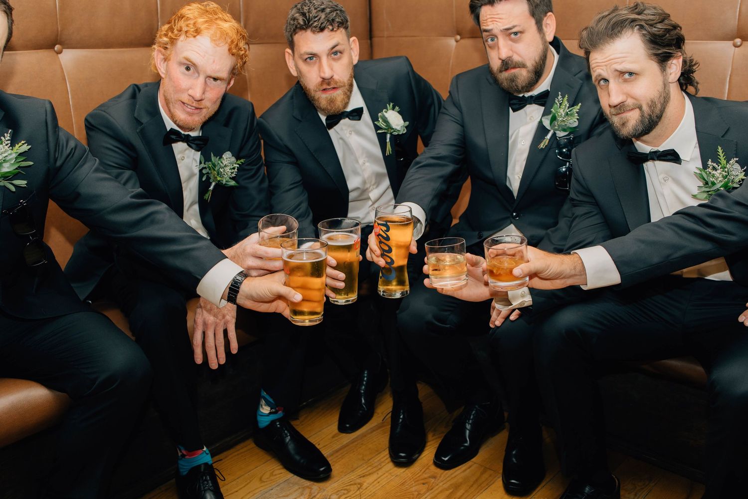 Four groomsmen in black tuxedos toast with beer glasses on a leather couch at a hotel in Paris Ontario.