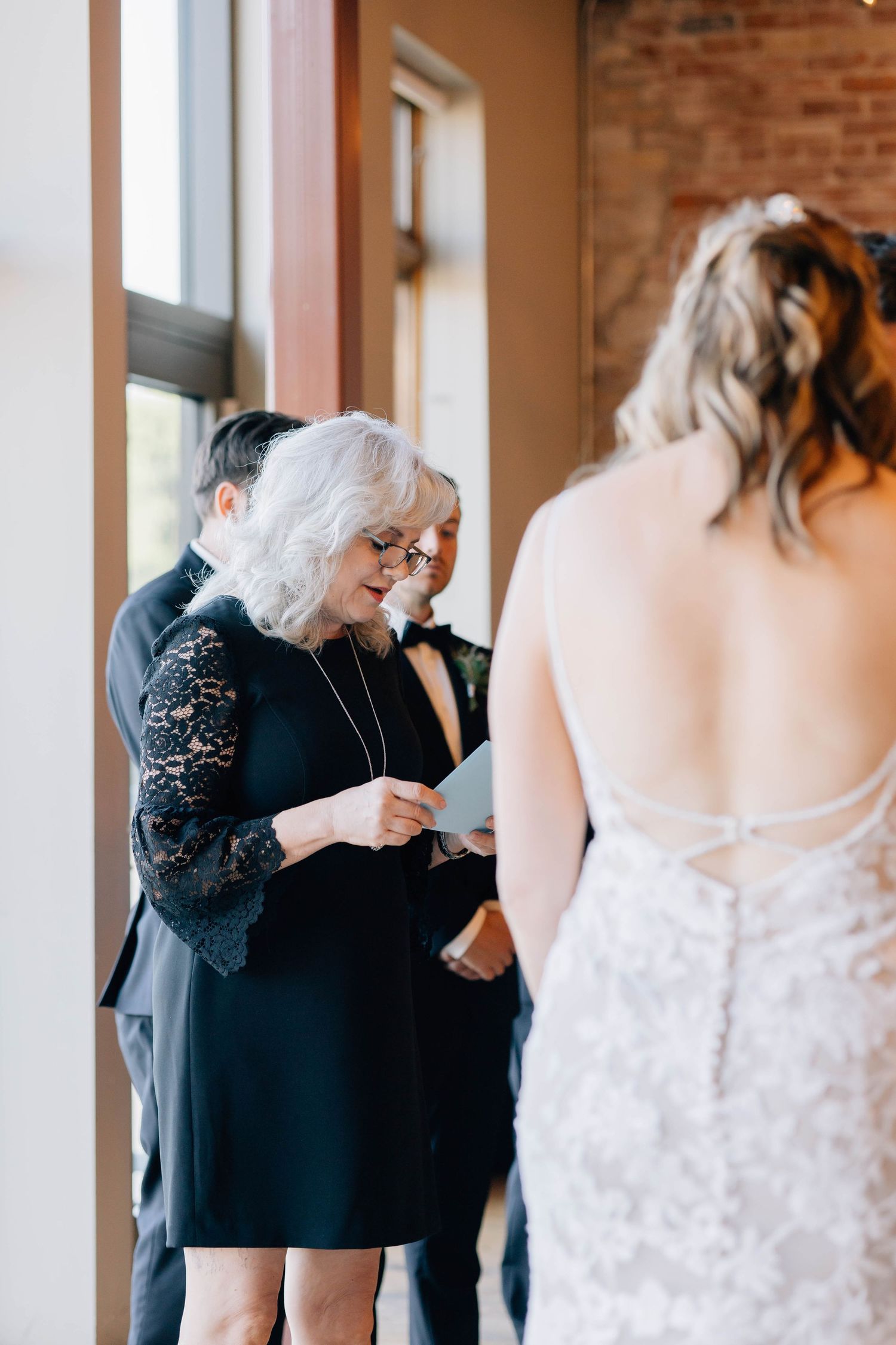 Elderly woman in black lace reads a card to the bride in a lace wedding gown during a pre-ceremony moment.