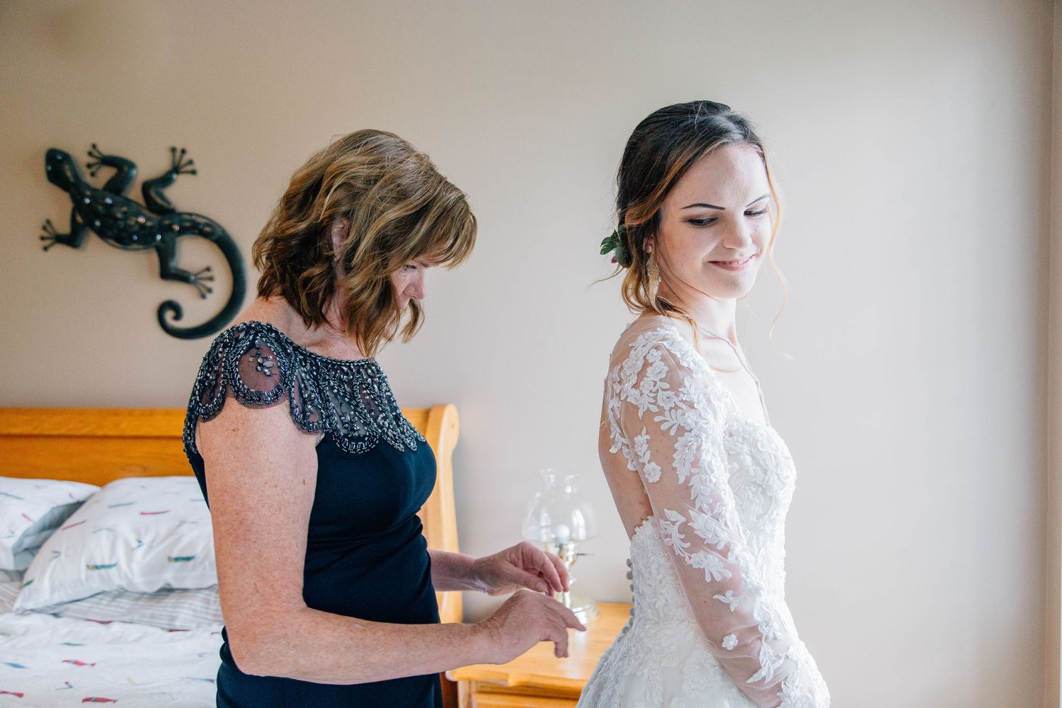 Mother helping bride fasten her elegant lace wedding gown in a warmly lit bedroom.
