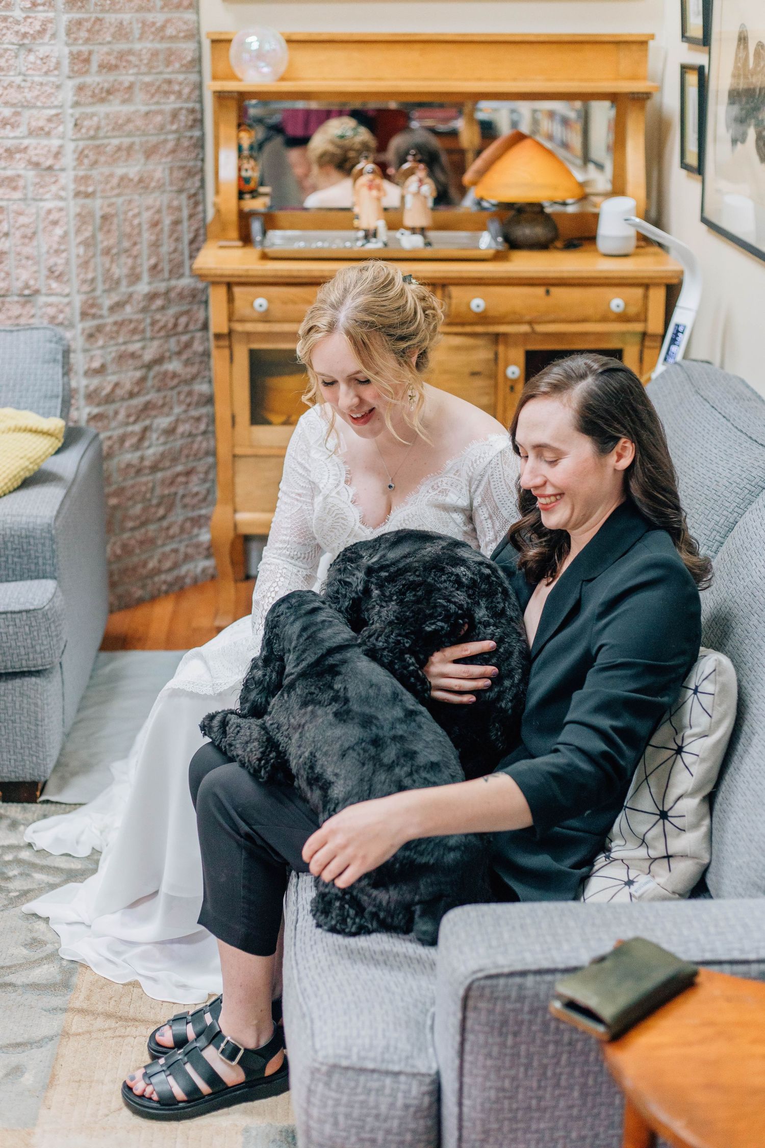 Bride and partner in black blazer cuddle large black dog together on gray sofa.
