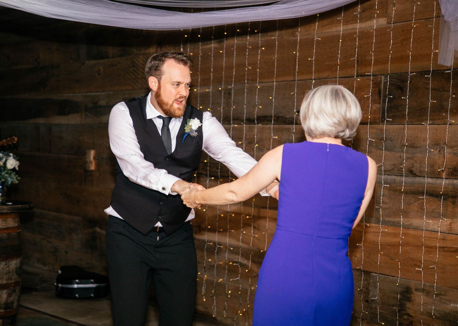 Groom dancing with mother in purple dress during mother-son dance at rustic barn wedding.