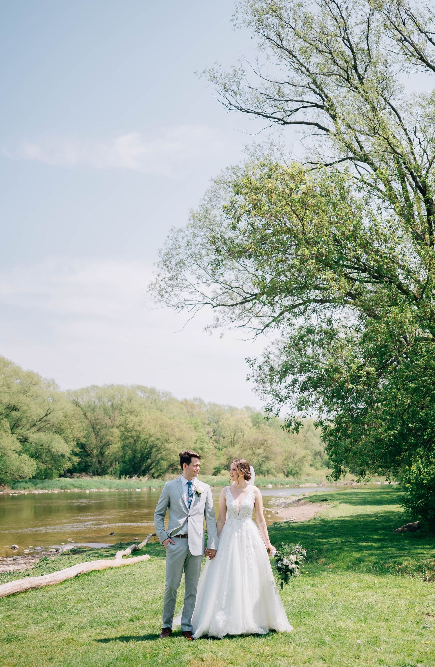 Bride and groom holding hands by a scenic riverside with lush green trees on a sunny spring wedding day.