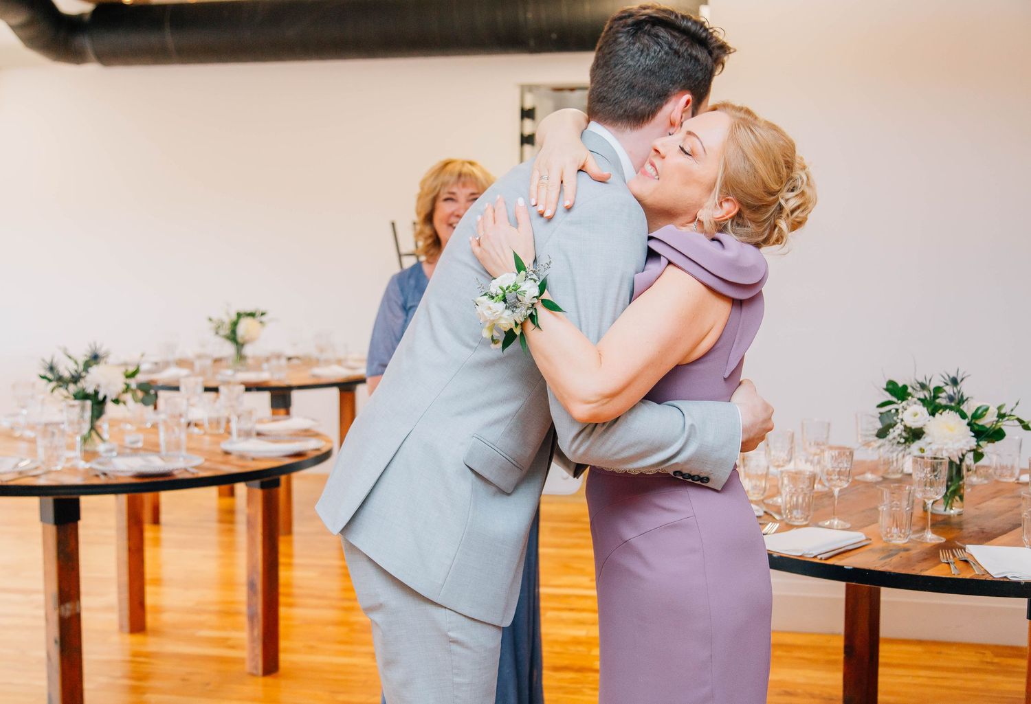 Groom in gray suit shares warm embrace with mother wearing purple dress at wedding reception.