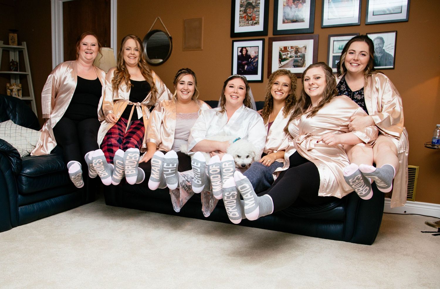 Bridal party in matching robes and white socks sitting on a couch laughing together during wedding morning preparations.