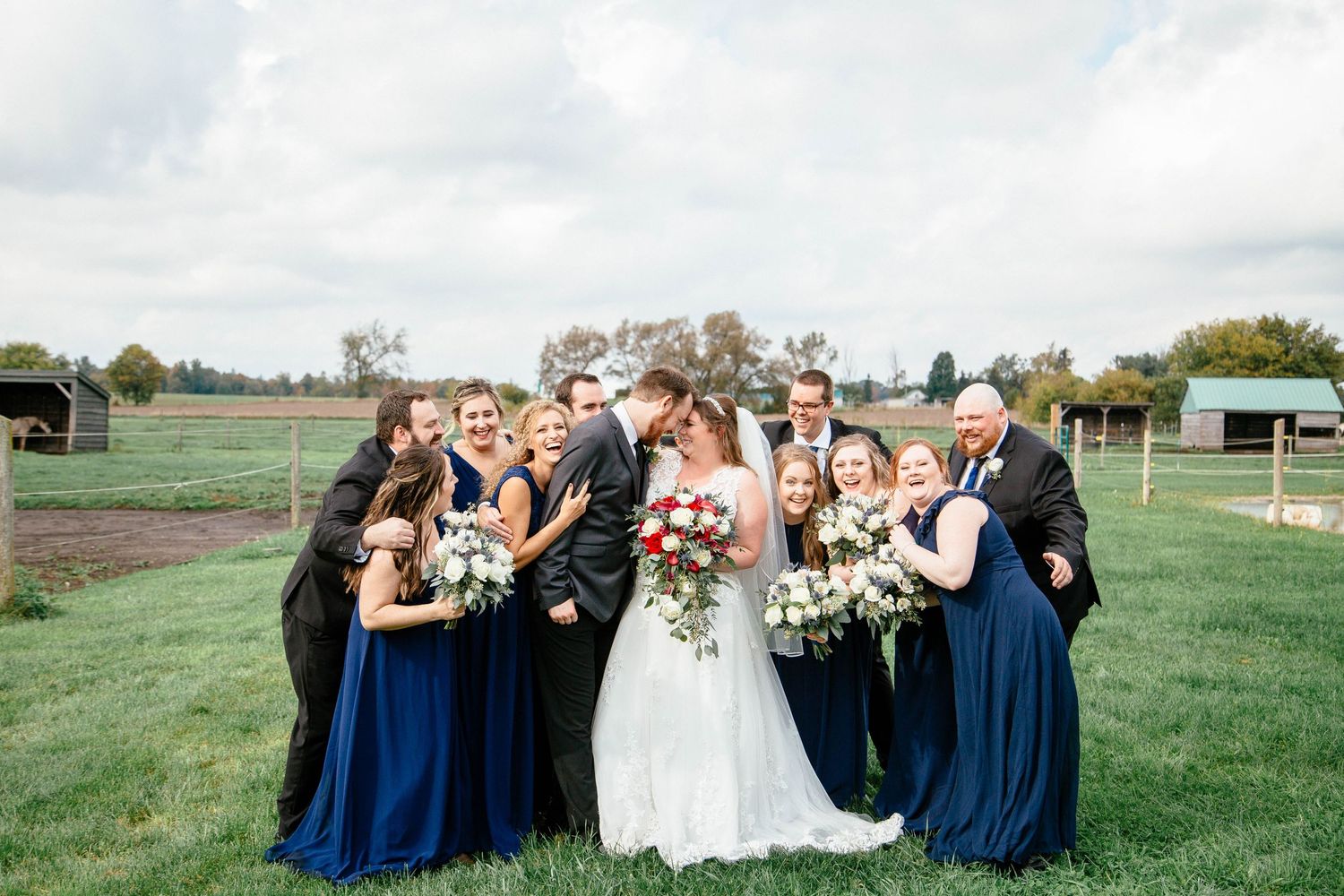 Wedding party surrounds bride and groom in a joyful group hug on a green farm lawn with bouquets of red and white flowers.
