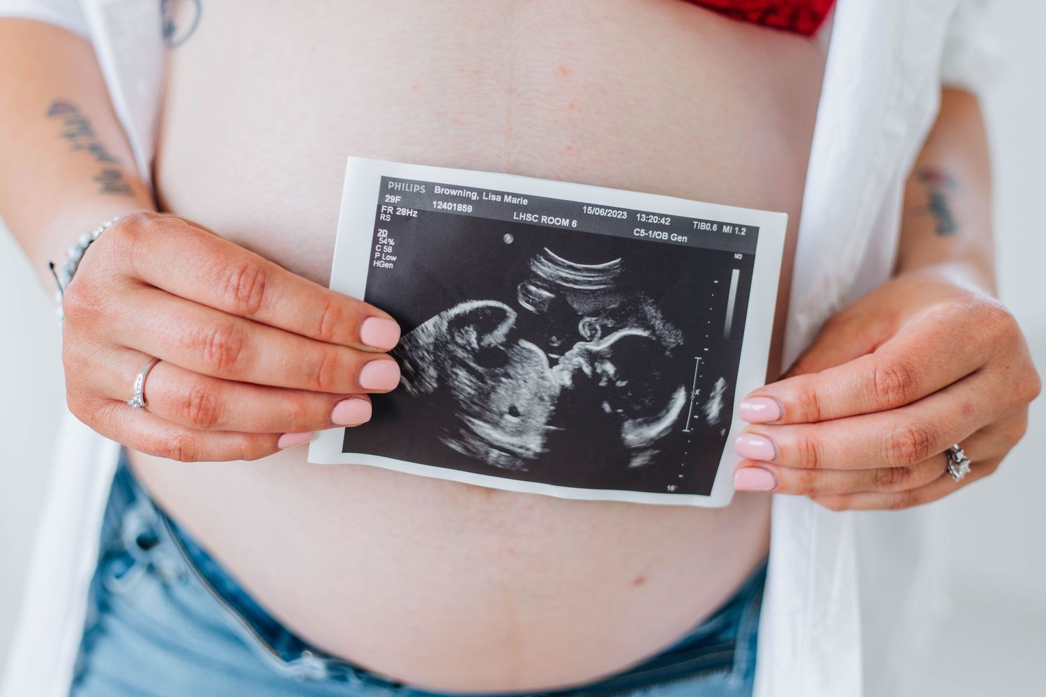 Close-up of pregnant belly with tattooed hands holding a baby ultrasound sonogram photo.