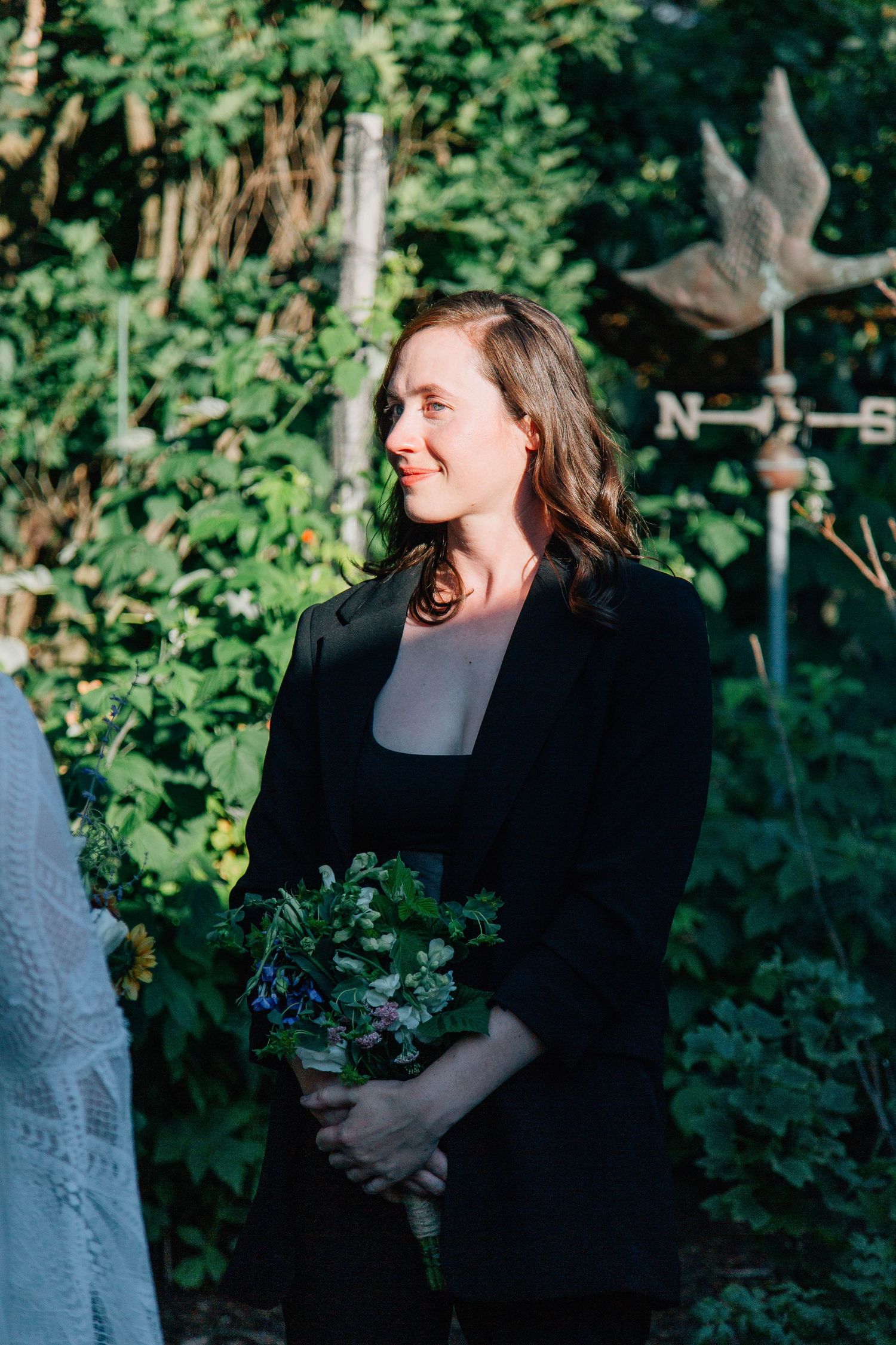 Woman in black blazer holds wildflower bouquet during outdoor wedding ceremony in green garden.