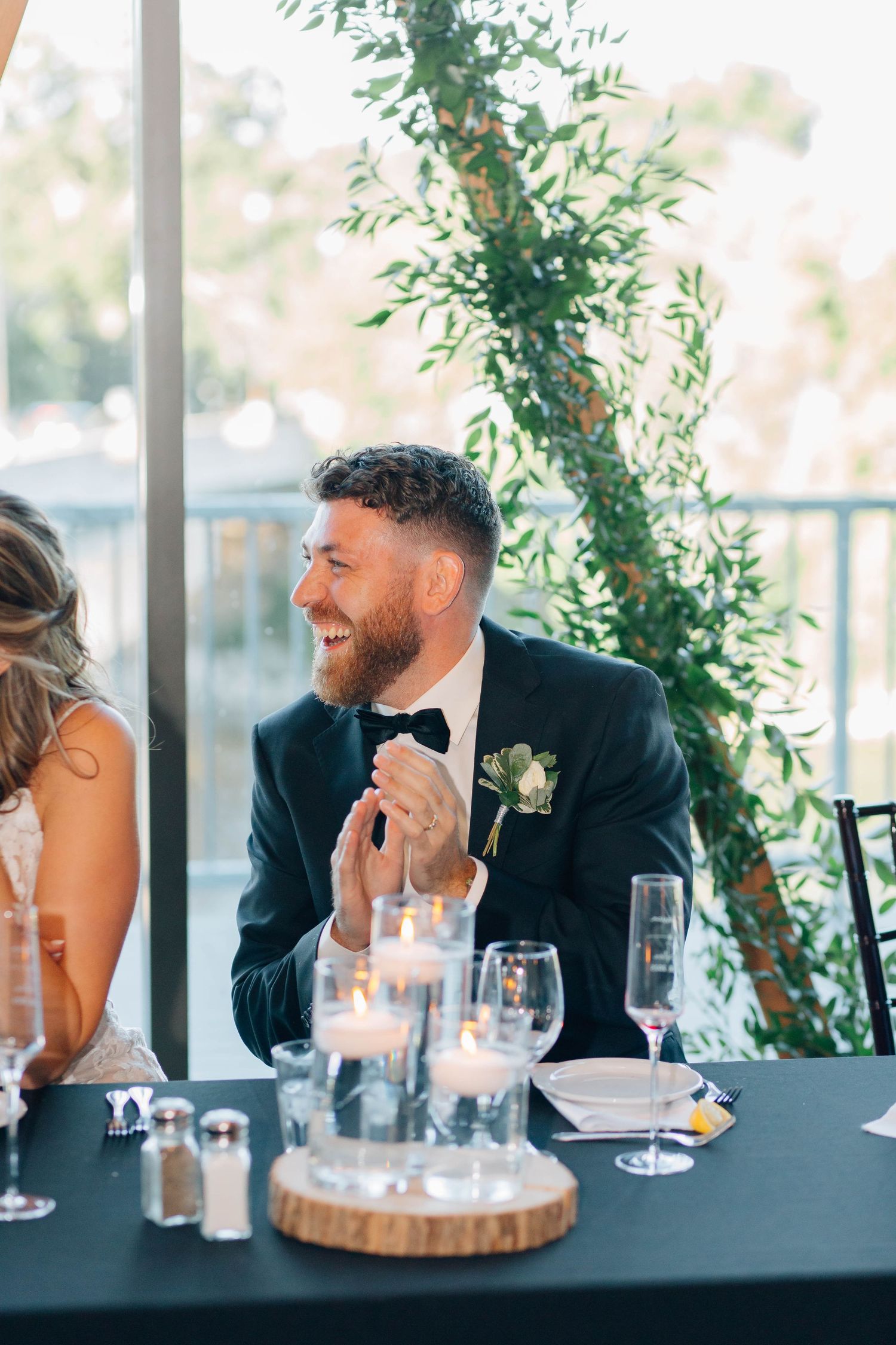 Laughing groom in black tuxedo with boutonniere sits at candlelit wedding reception table with greenery backdrop.