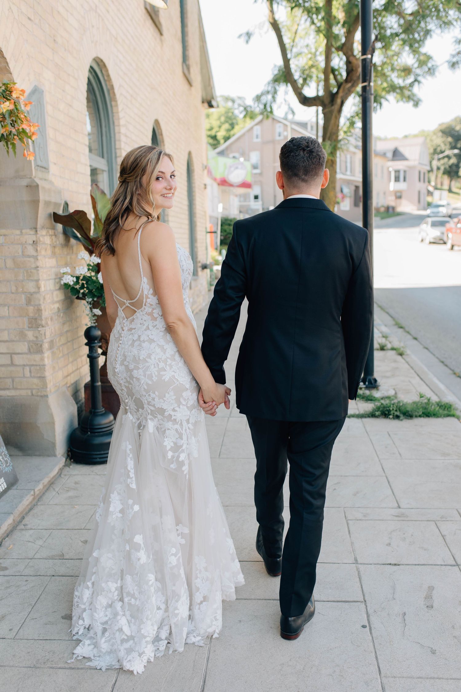 Bride in lace backless gown holds hands with groom in black tuxedo walking along sunny sidewalk in Paris Ontario.