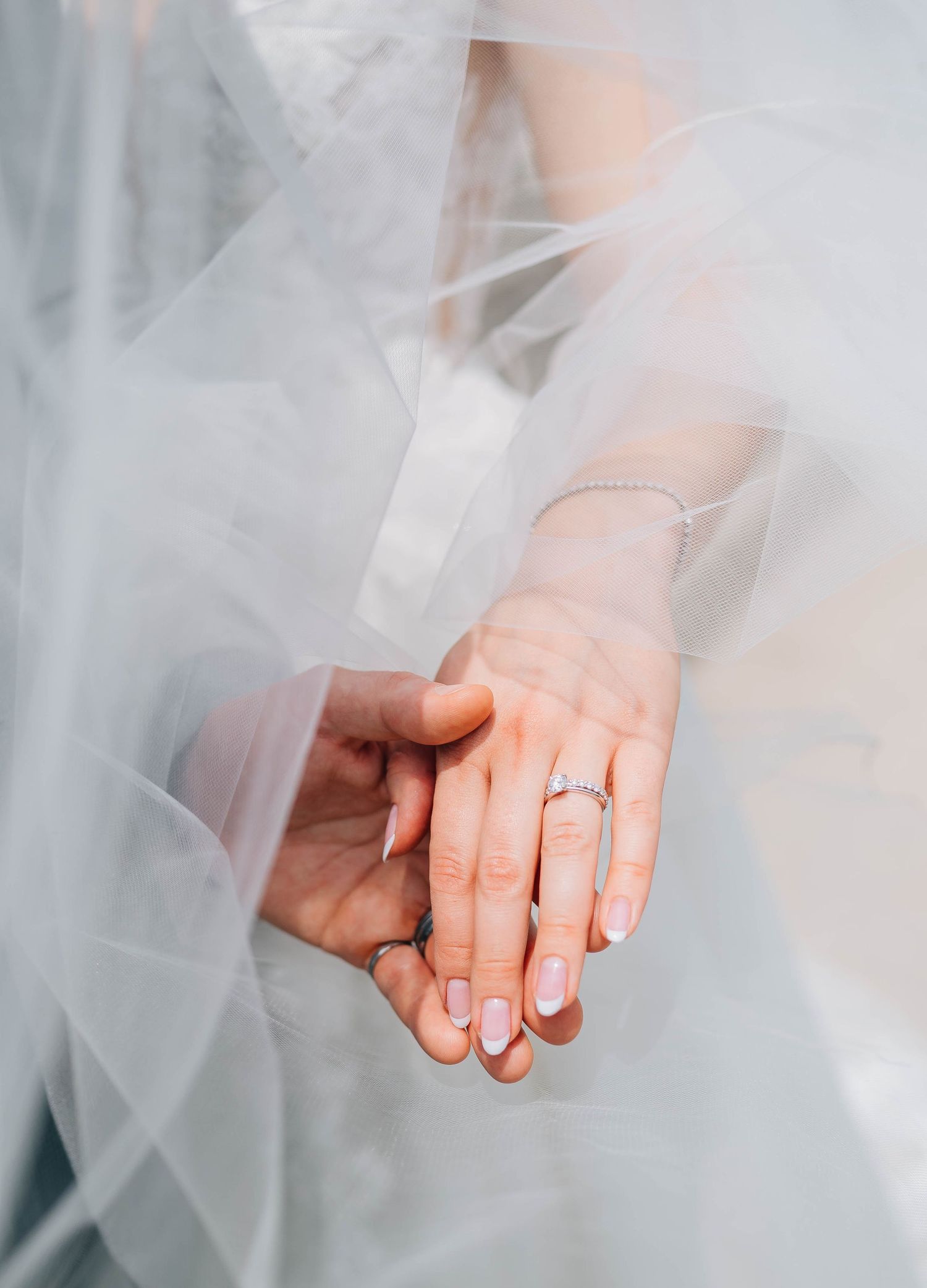 Close-up of bride and groom holding hands, showcasing wedding ring and veil.
