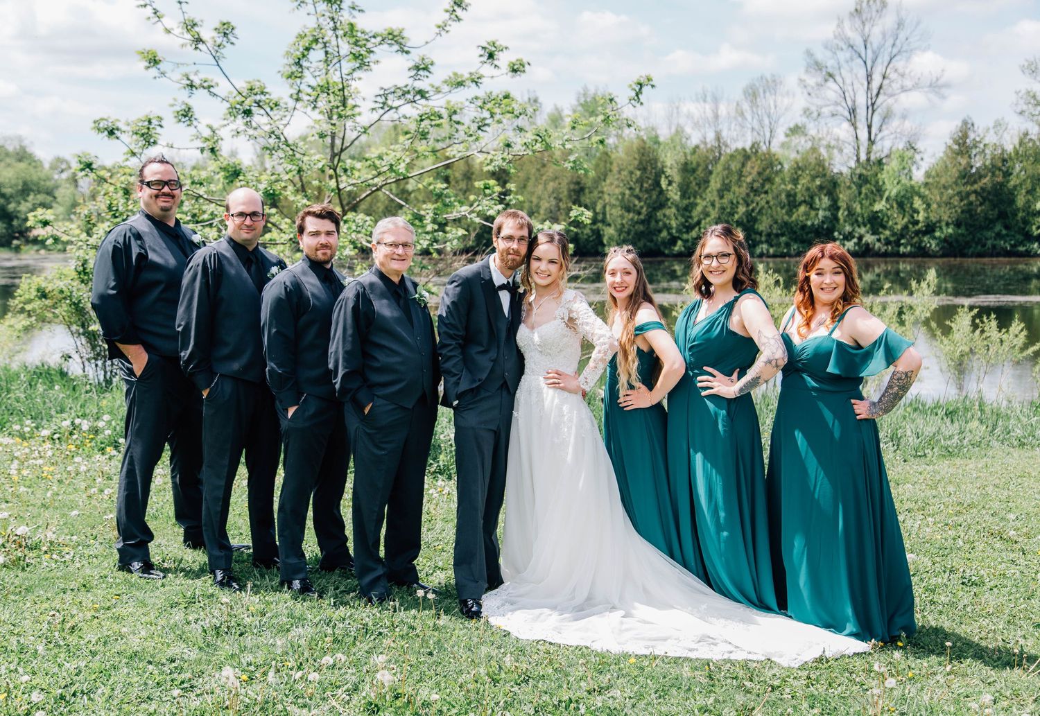 Wedding party posing together outdoors, bridesmaids in teal gowns and groomsmen in black suits by a pond.