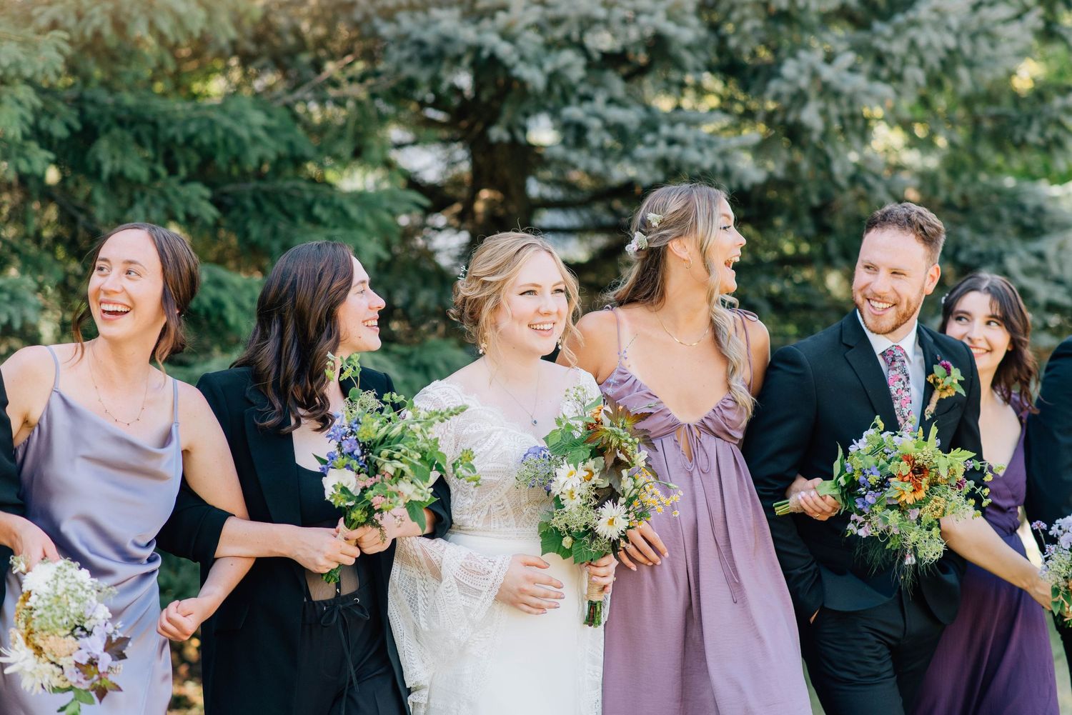 Bride laughing with bridesmaids in purple dresses holding wildflower bouquets outdoors near evergreen trees.