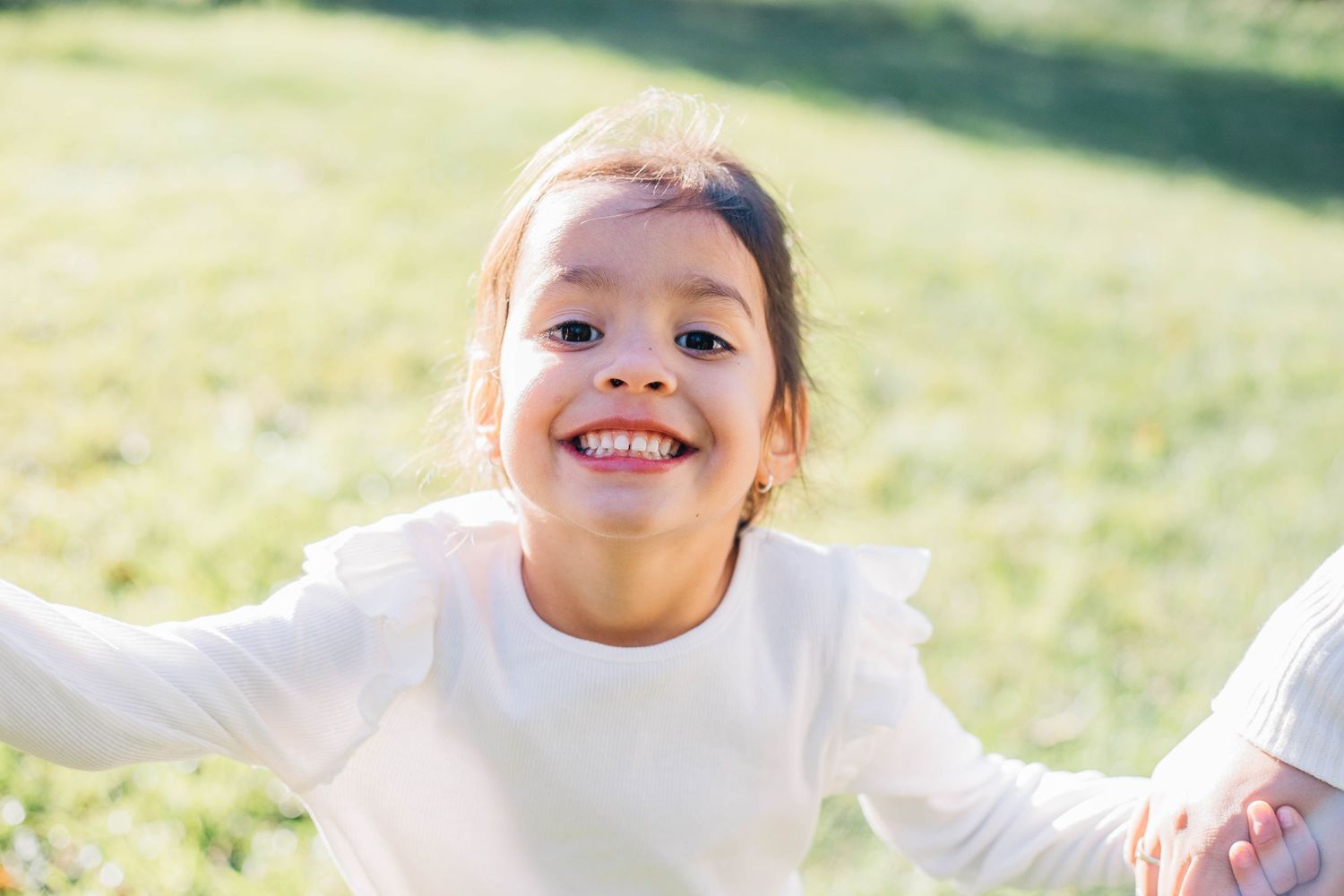 Adorable young girl in white top smiling broadly with arms outstretched in a sunny green field.