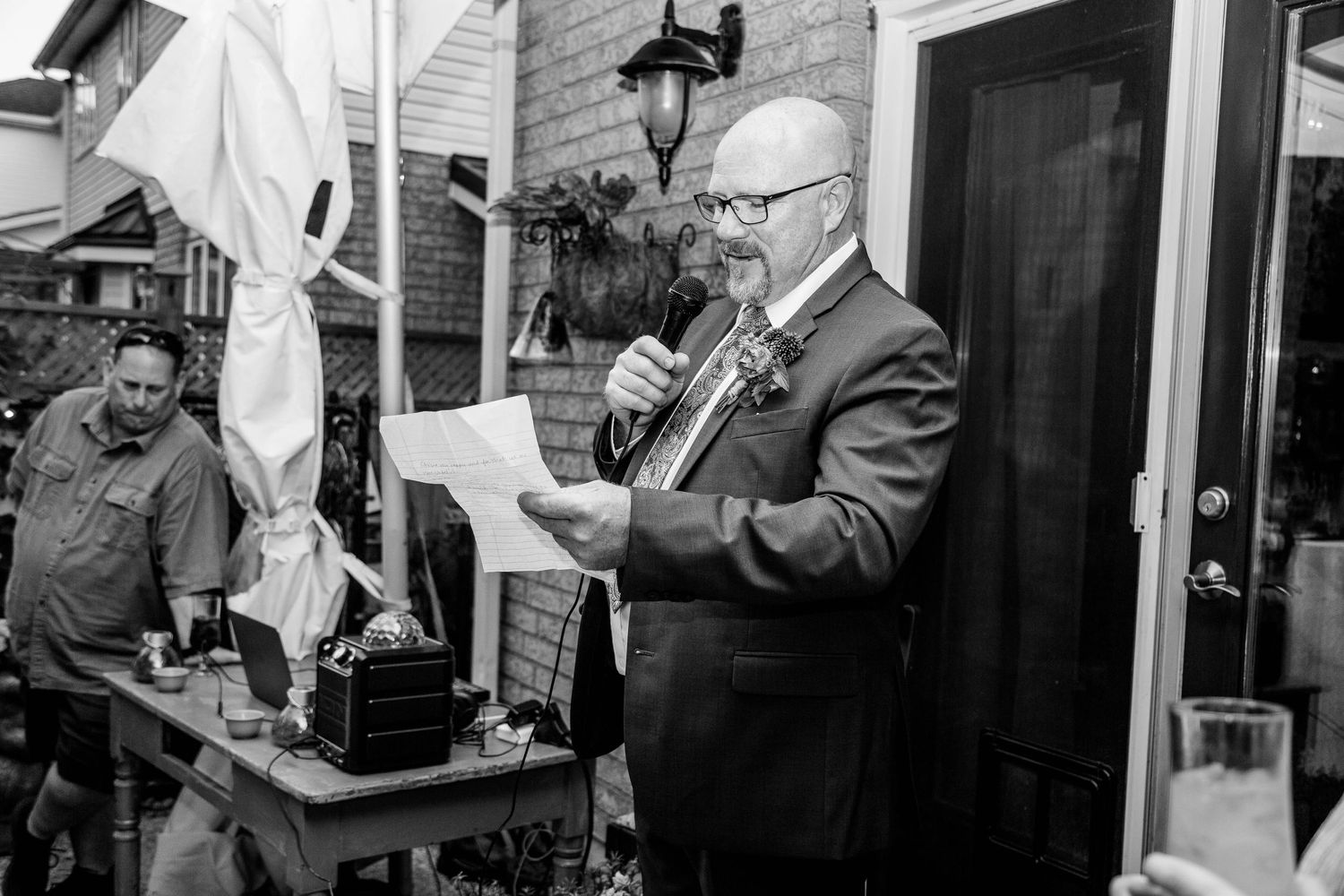 Man in suit reads from paper during outdoor wedding speech in black and white photo.