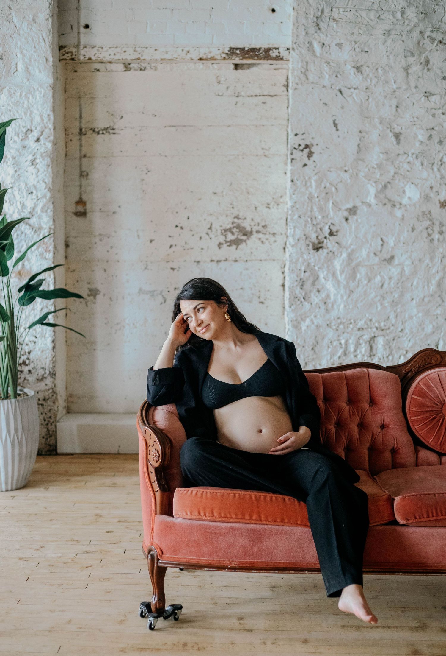 Pregnant woman in black blazer and bra lounging on a coral velvet sofa in a rustic loft studio.