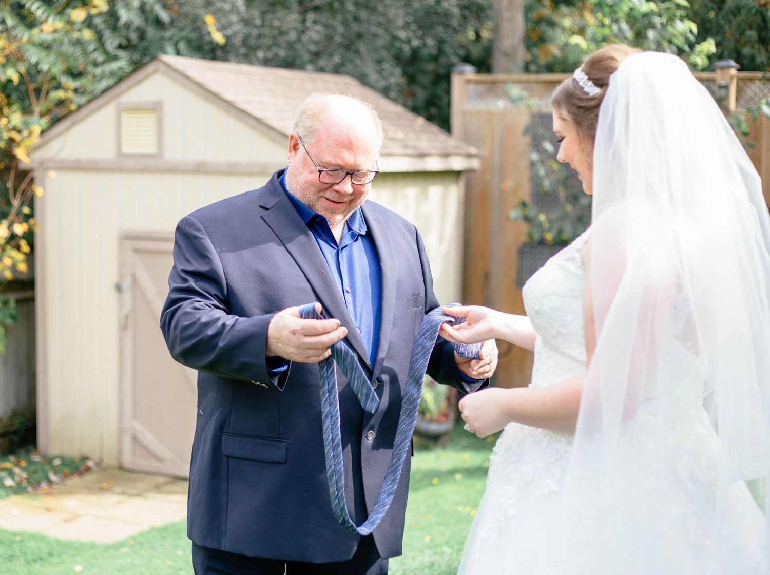 Bride sharing a tender moment with older man in navy suit during backyard wedding preparation.