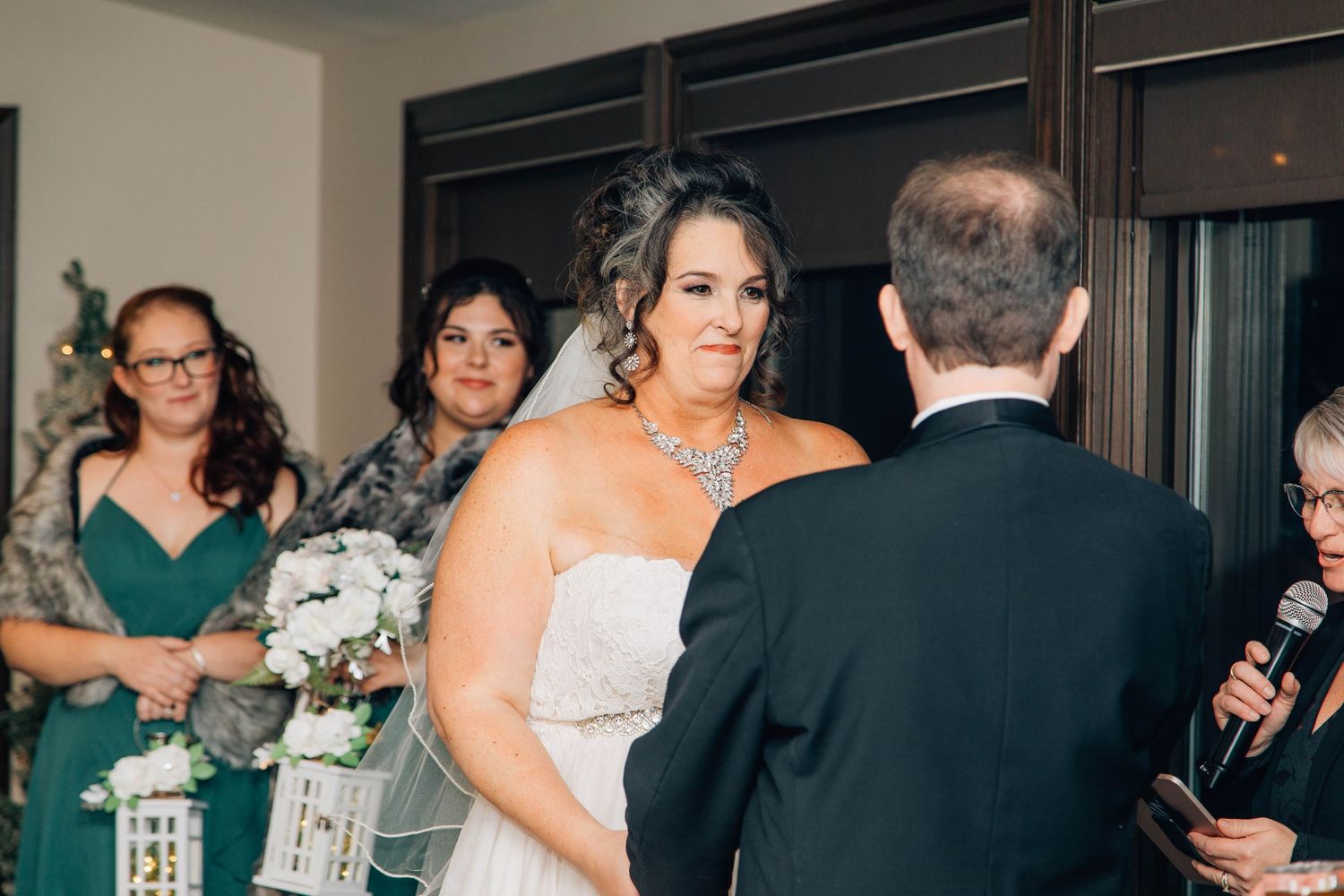 Bride in strapless gown with crystal necklace exchanges vows while bridesmaids in emerald green stand behind her.