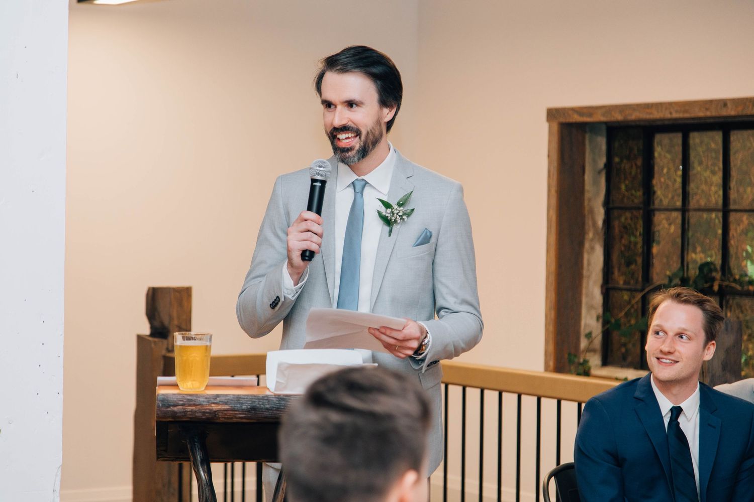 Groomsman in gray suit delivering a heartfelt wedding speech with microphone at reception.