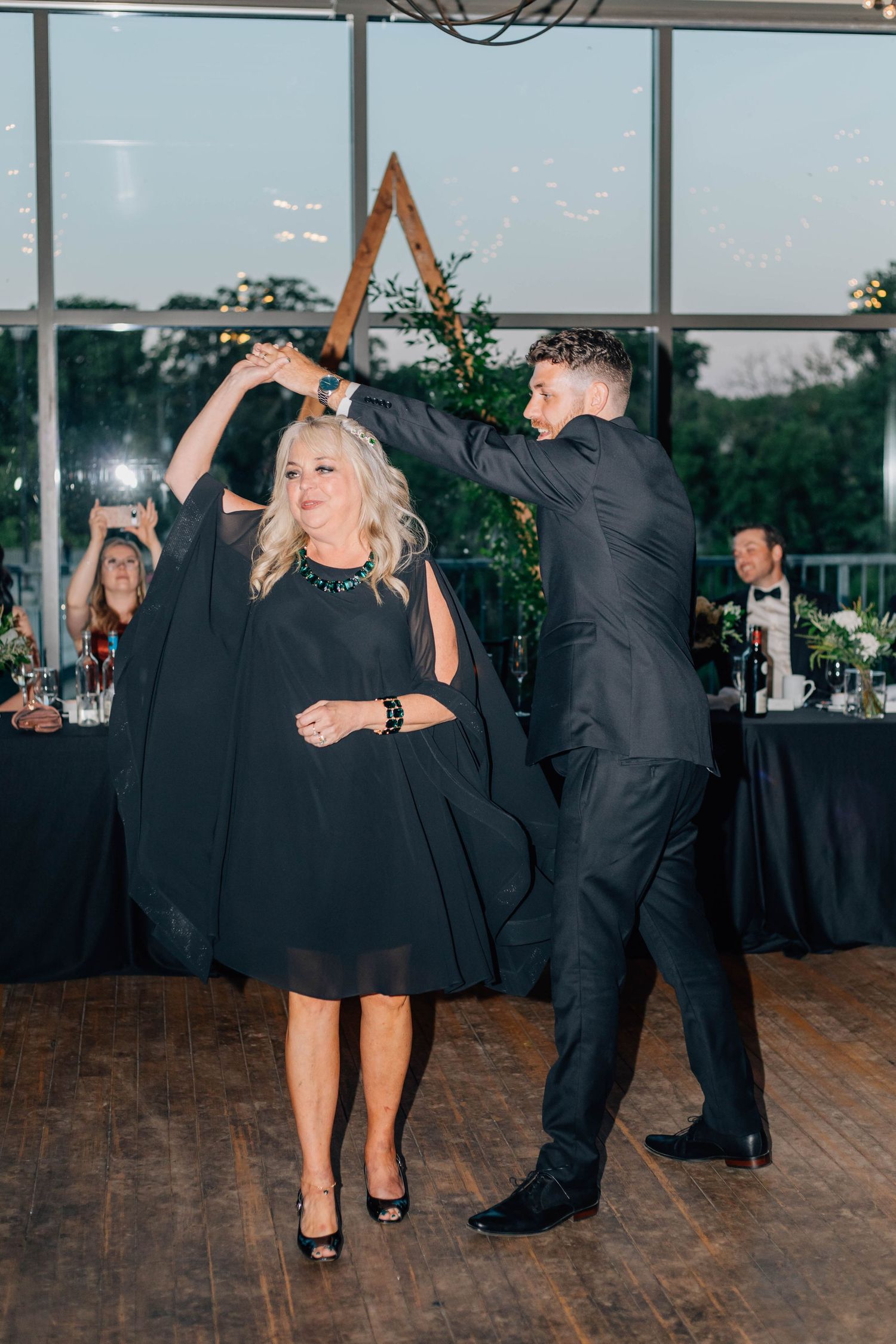 Groom in black tuxedo spins mother in black dress during mother-son dance at wedding reception.