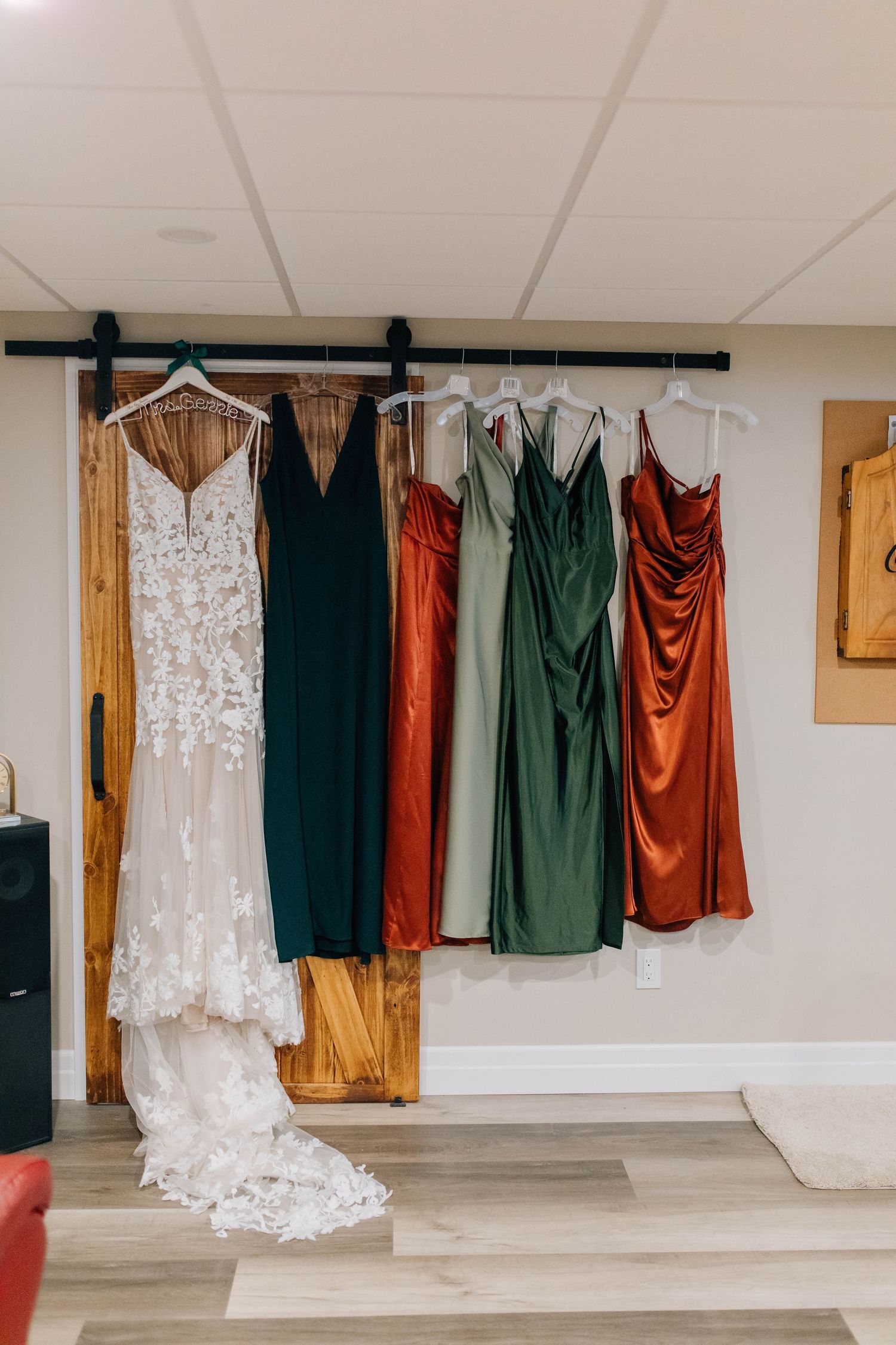 Wedding dress and colorful bridesmaid dresses in green, rust, and gold hanging on a barn door rack.