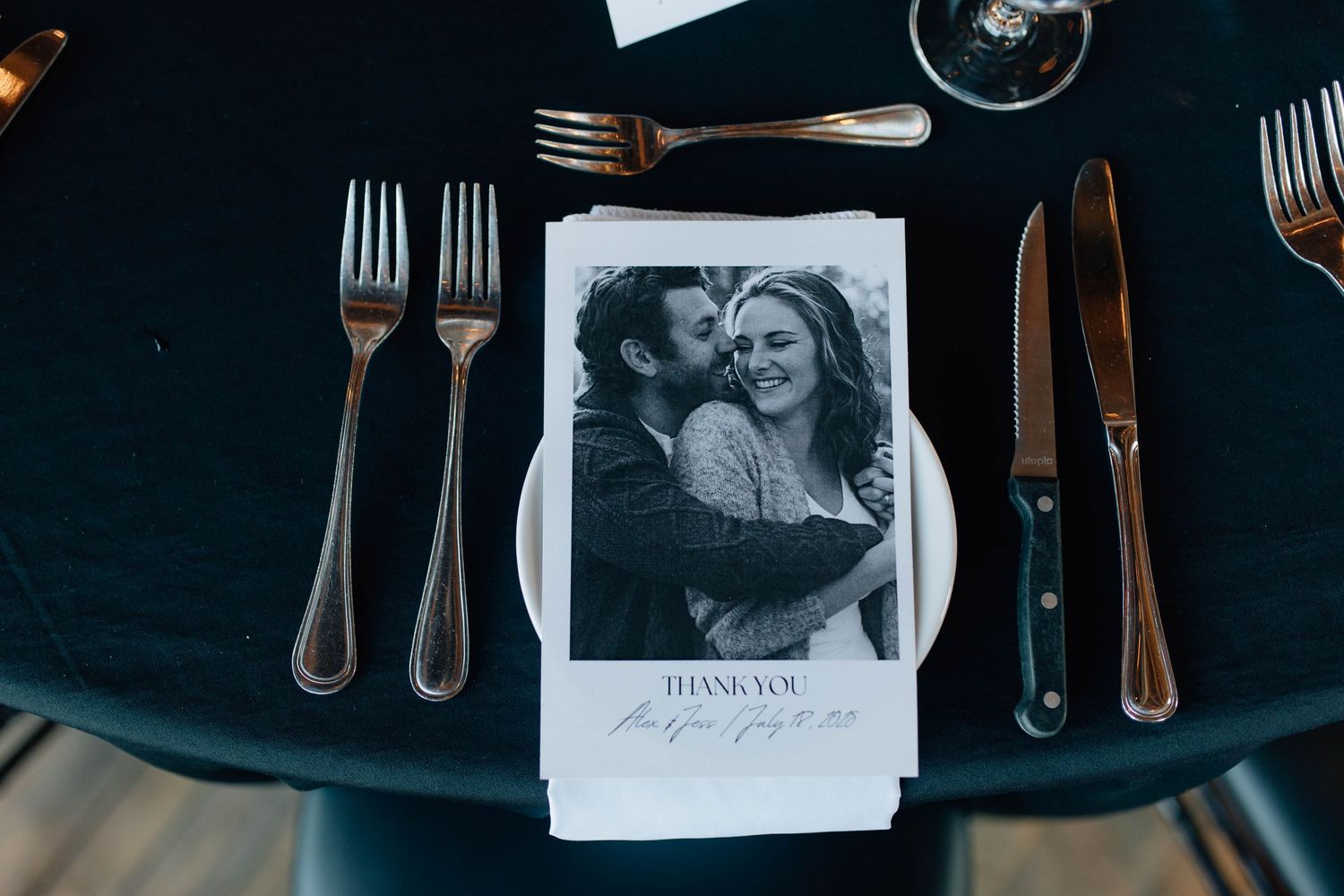 Wedding place setting featuring black-and-white thank you card with couple photo, gold cutlery on black tablecloth.