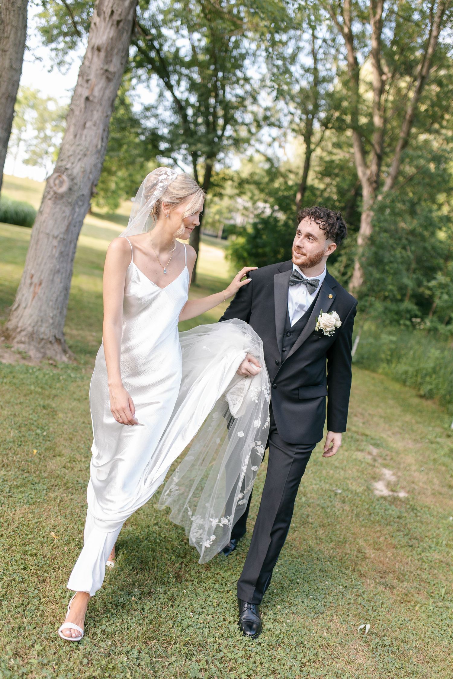Bride in white satin gown and groom in black tuxedo walking outdoors on grass with trees in background at Sydenham Ridge in Strathroy.