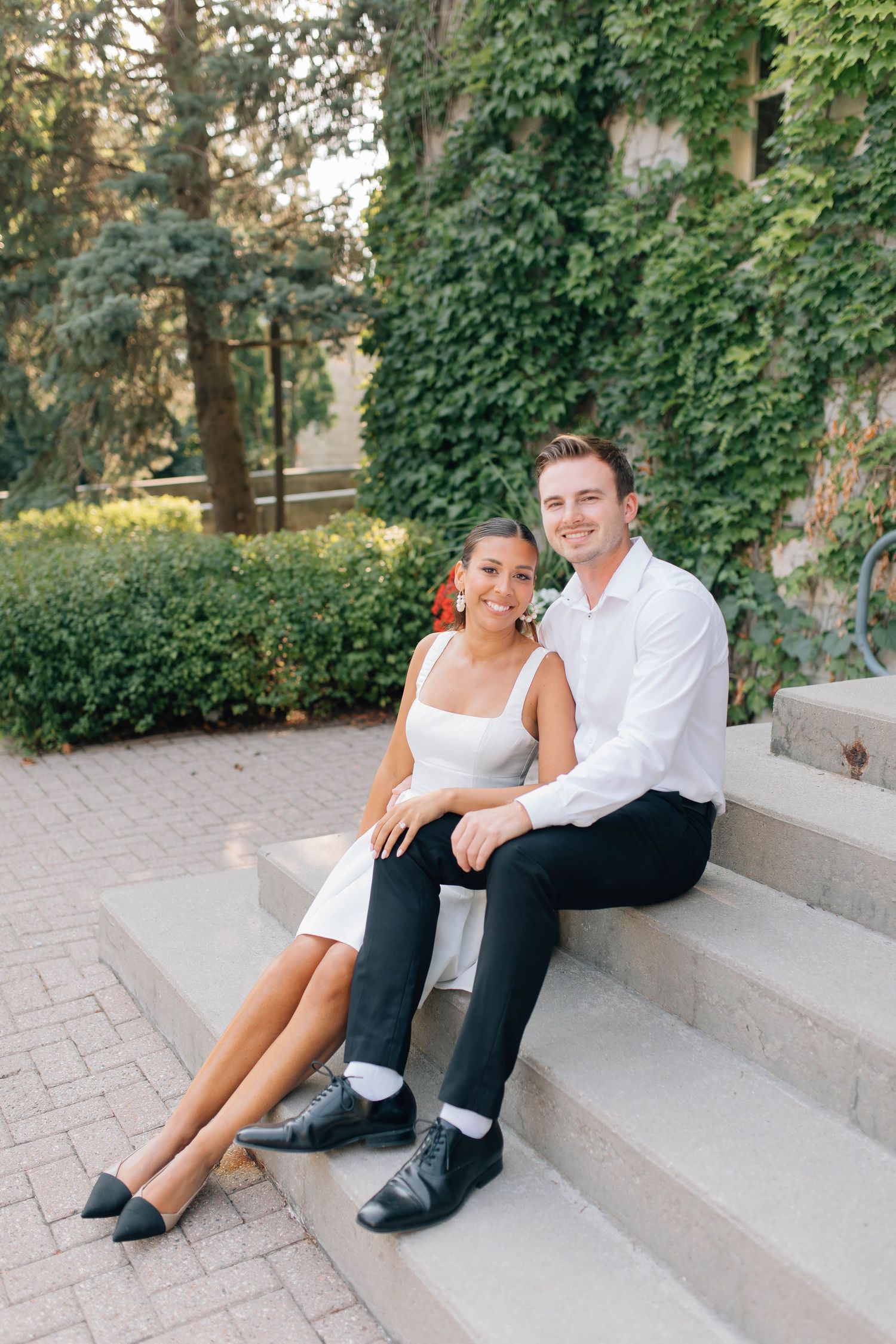 Engaged couple sits smiling together on stone steps outside ivy-covered building in white and black outfits.