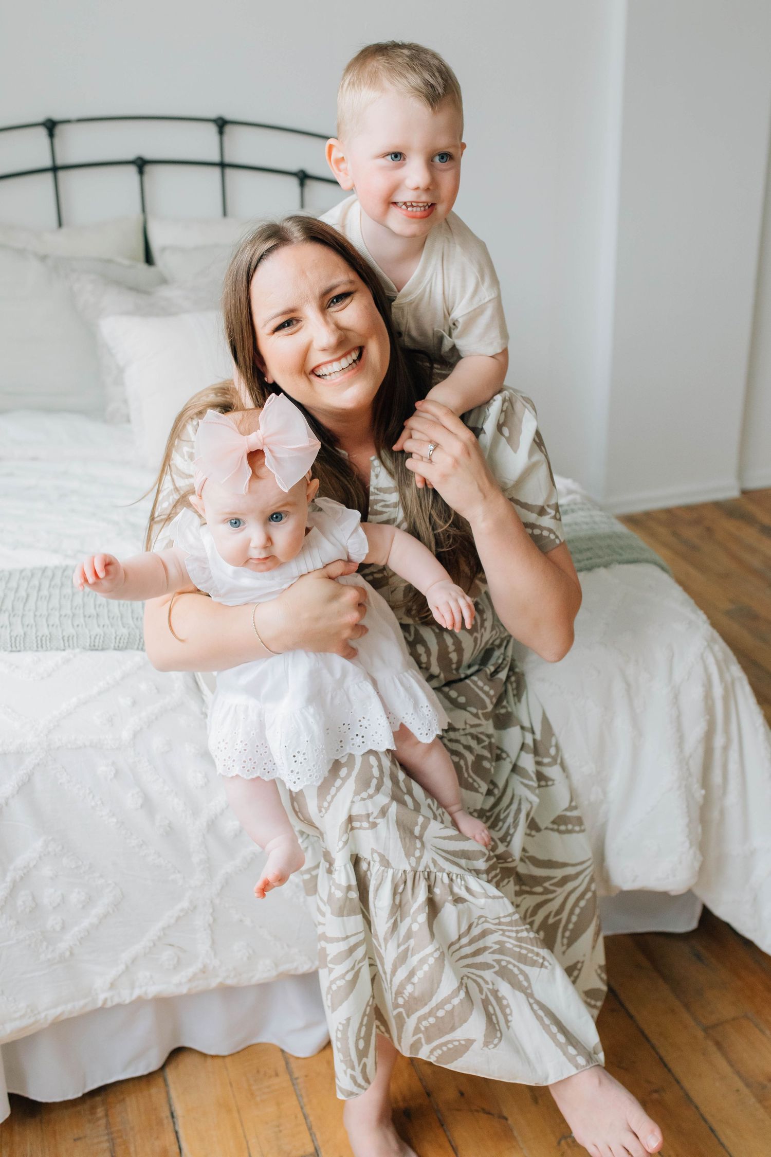 Smiling mom sitting on bed holding baby girl in white dress while toddler boy hugs her from behind.