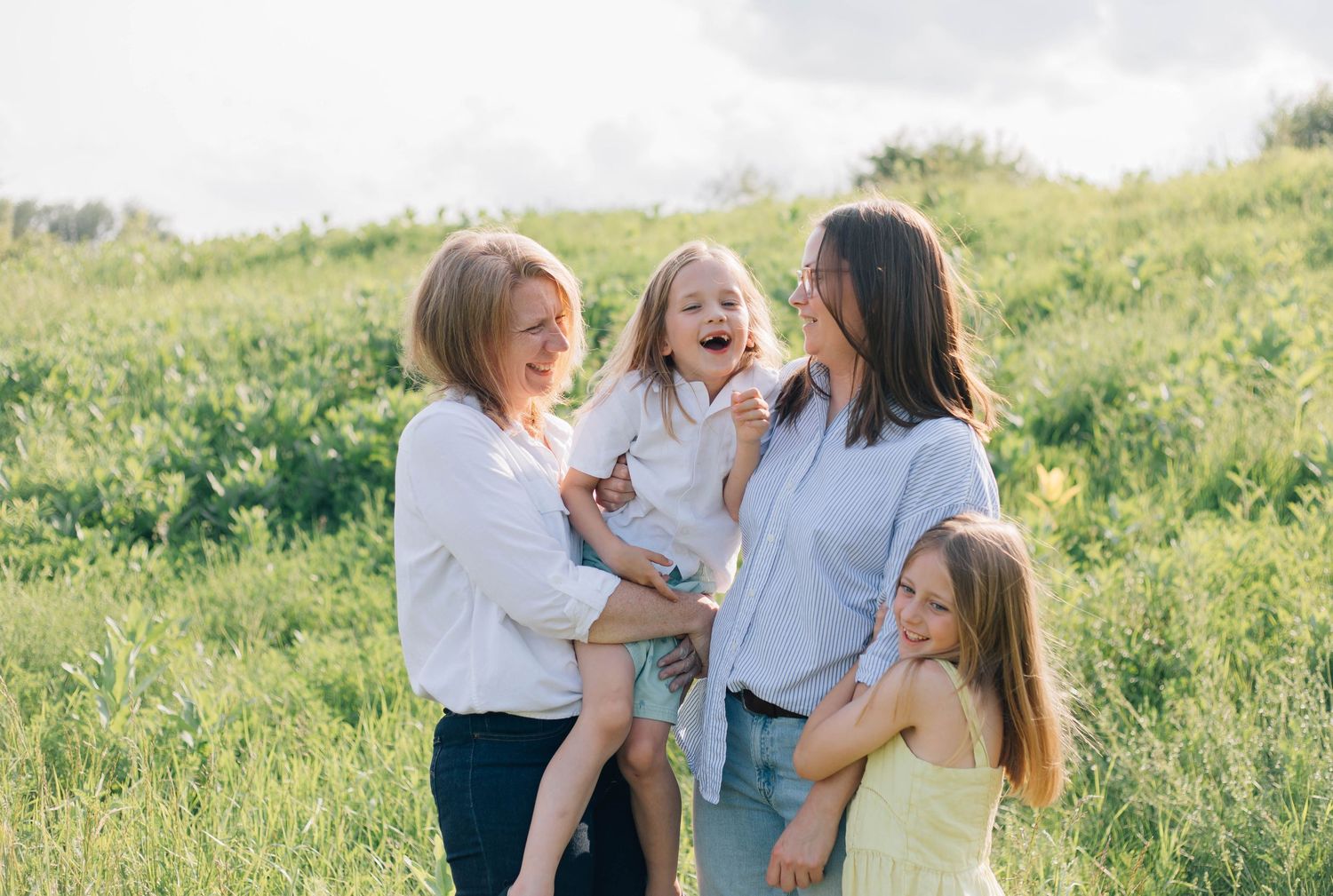 Two moms laughing outdoors in green field, one holding toddler while older daughter hugs her side.