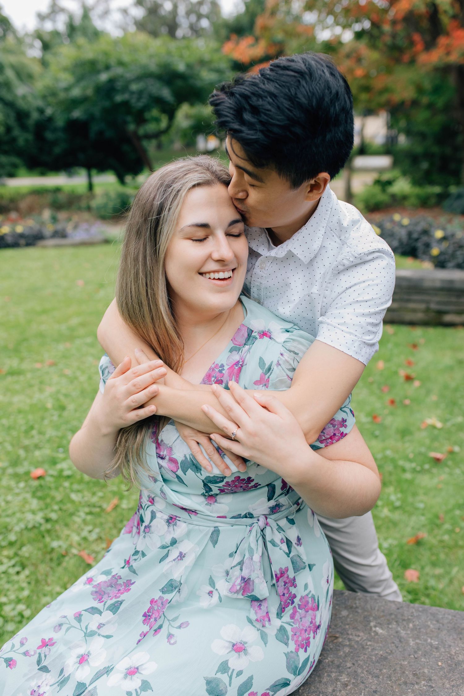 Man embracing woman from behind and kissing her cheek as she smiles in a green park.