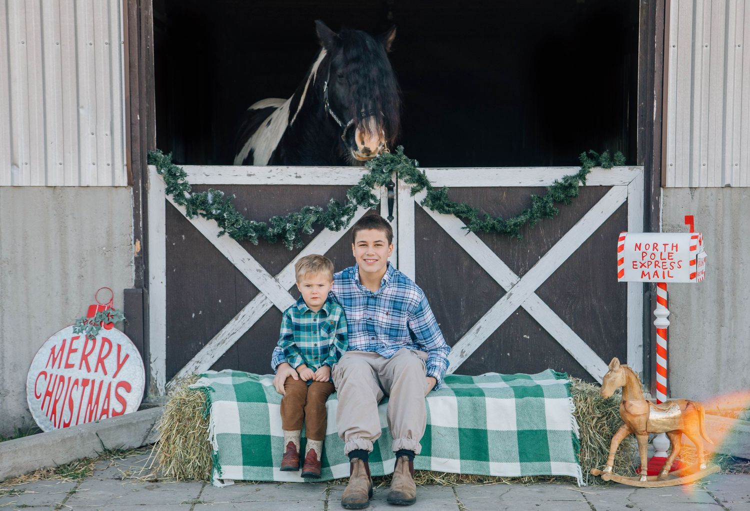 Two brothers sitting on hay bale with horse peeking from decorated Christmas barn door.