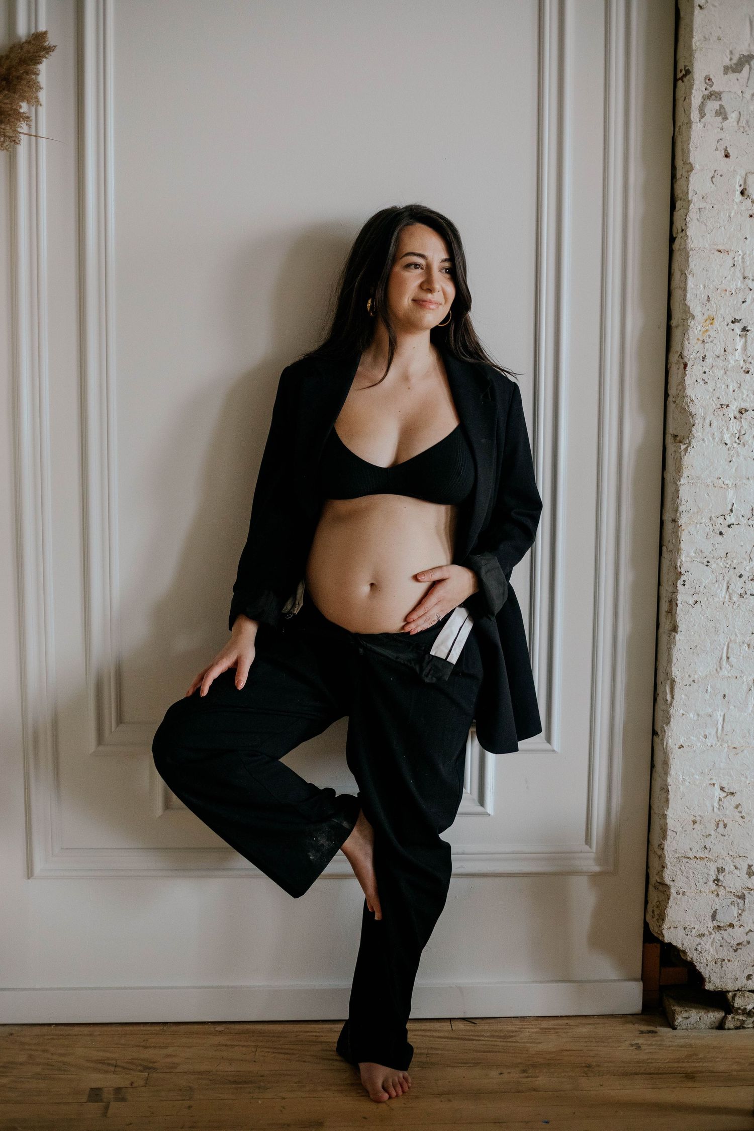 Pregnant woman in black blazer and bra posing confidently against a white wall with one leg raised.