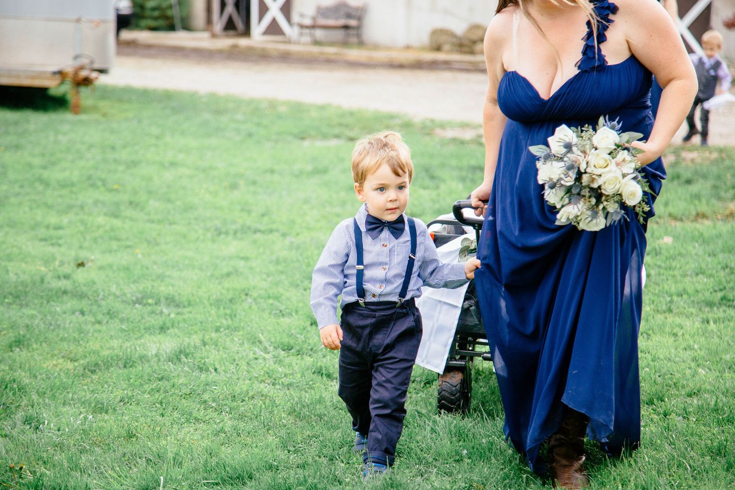 Young ring bearer in suspenders and a bridesmaid in a navy blue gown walk together across a green lawn.