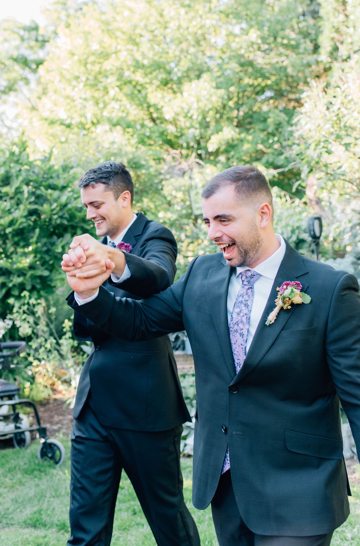 Two grooms in dark suits laugh and celebrate together outdoors after wedding ceremony.