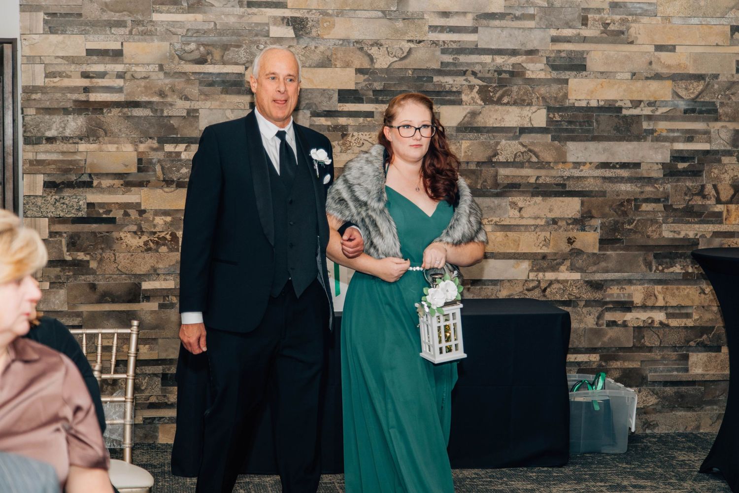 Bridesmaid in emerald green gown with fur wrap walks down aisle with groomsman at winter wedding reception.