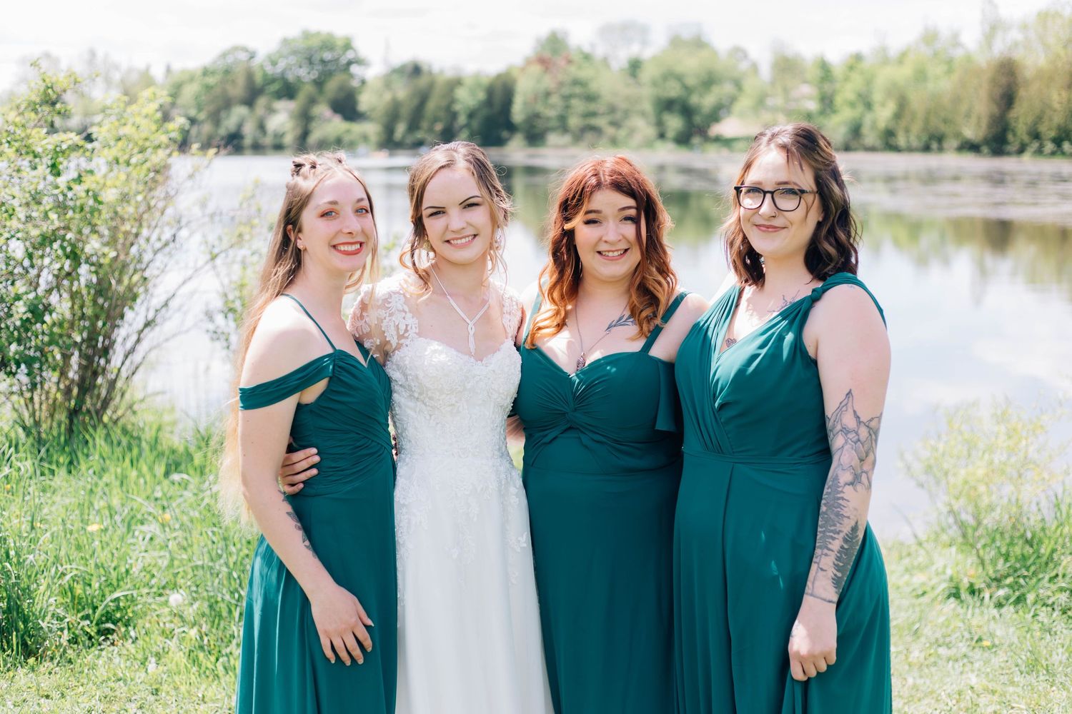 Bride in lace gown posing with three bridesmaids in emerald green dresses by a lake.