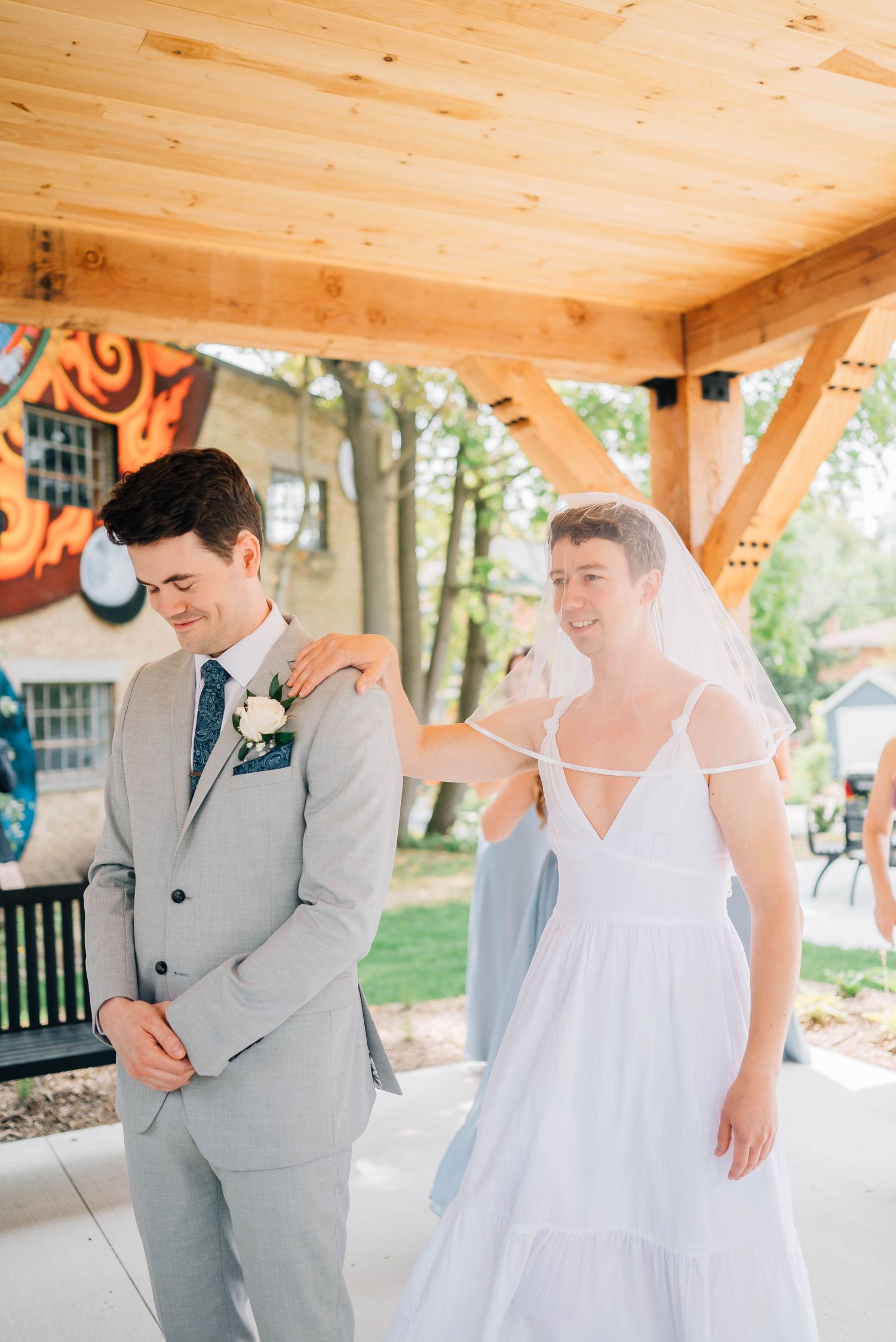 Bride in white dress and veil taps groom on shoulder during outdoor first look.