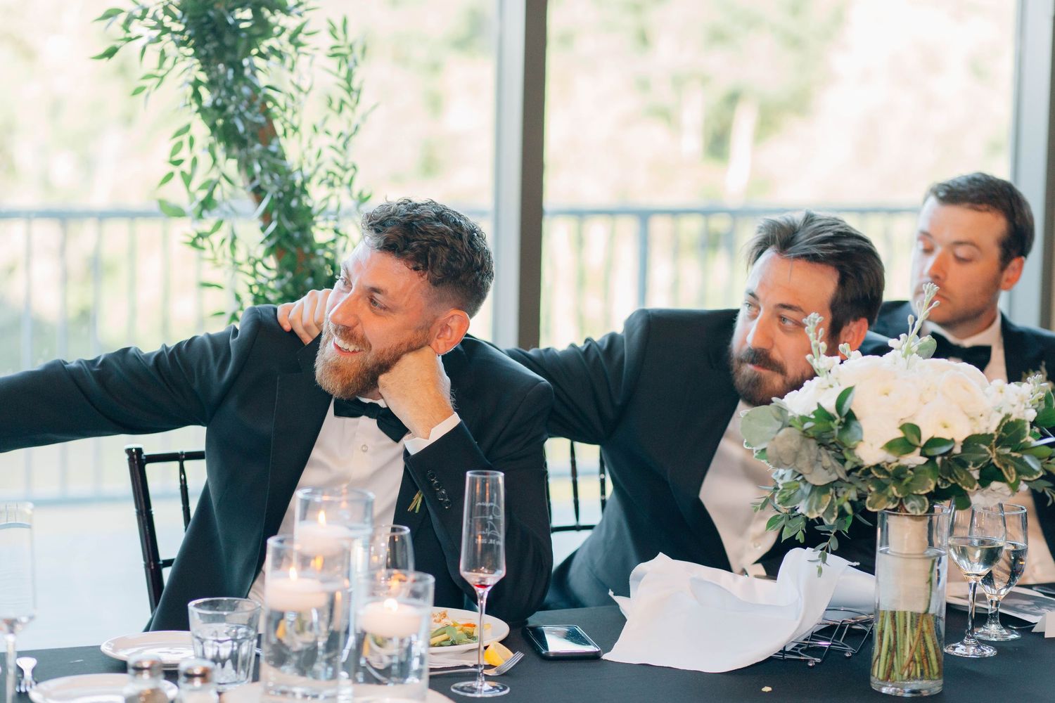 Two groomsmen in black tuxedos laugh together at wedding reception head table with floral bouquet.