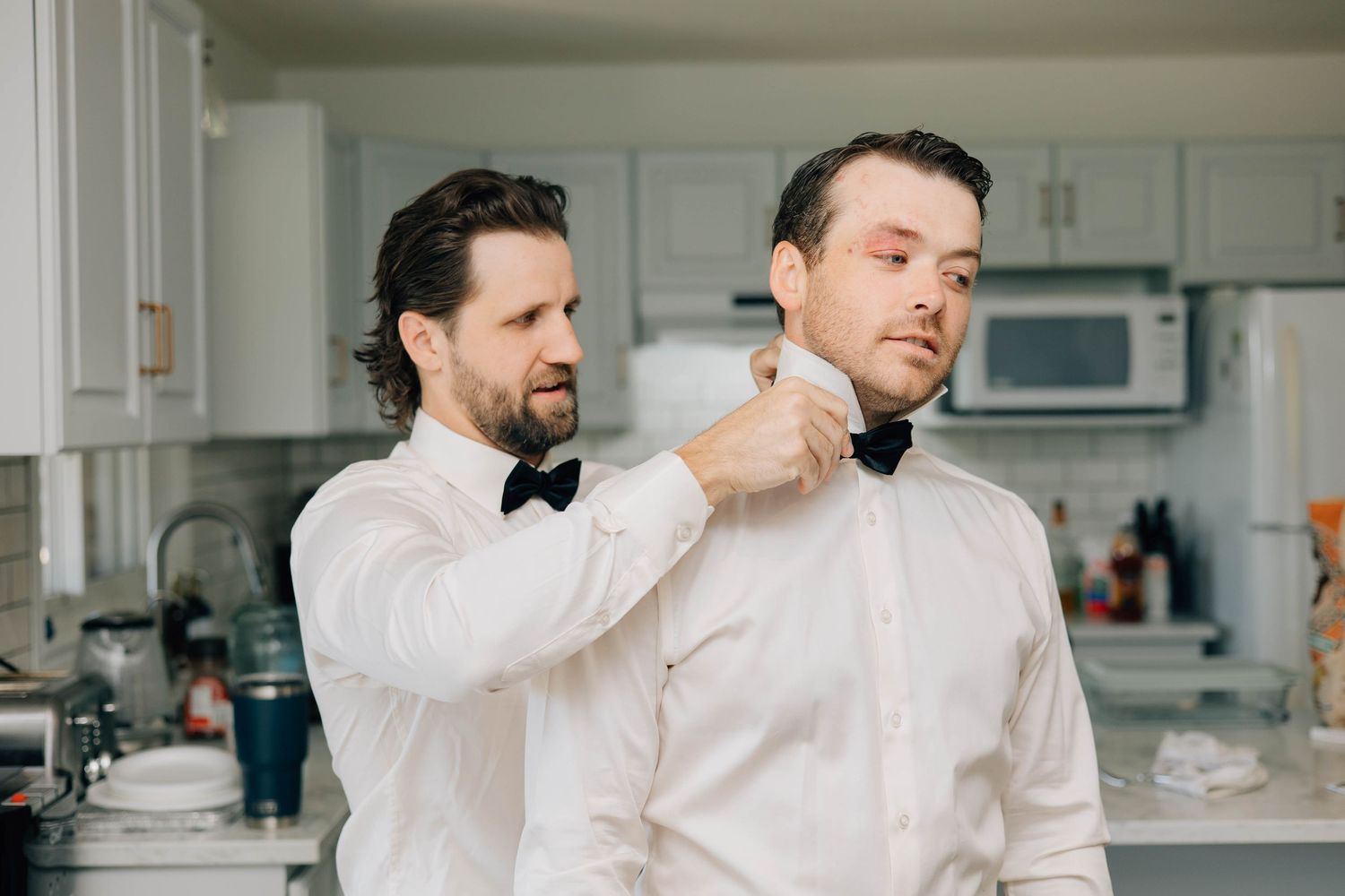 Man helping another adjust black bow tie in a bright kitchen before wedding.