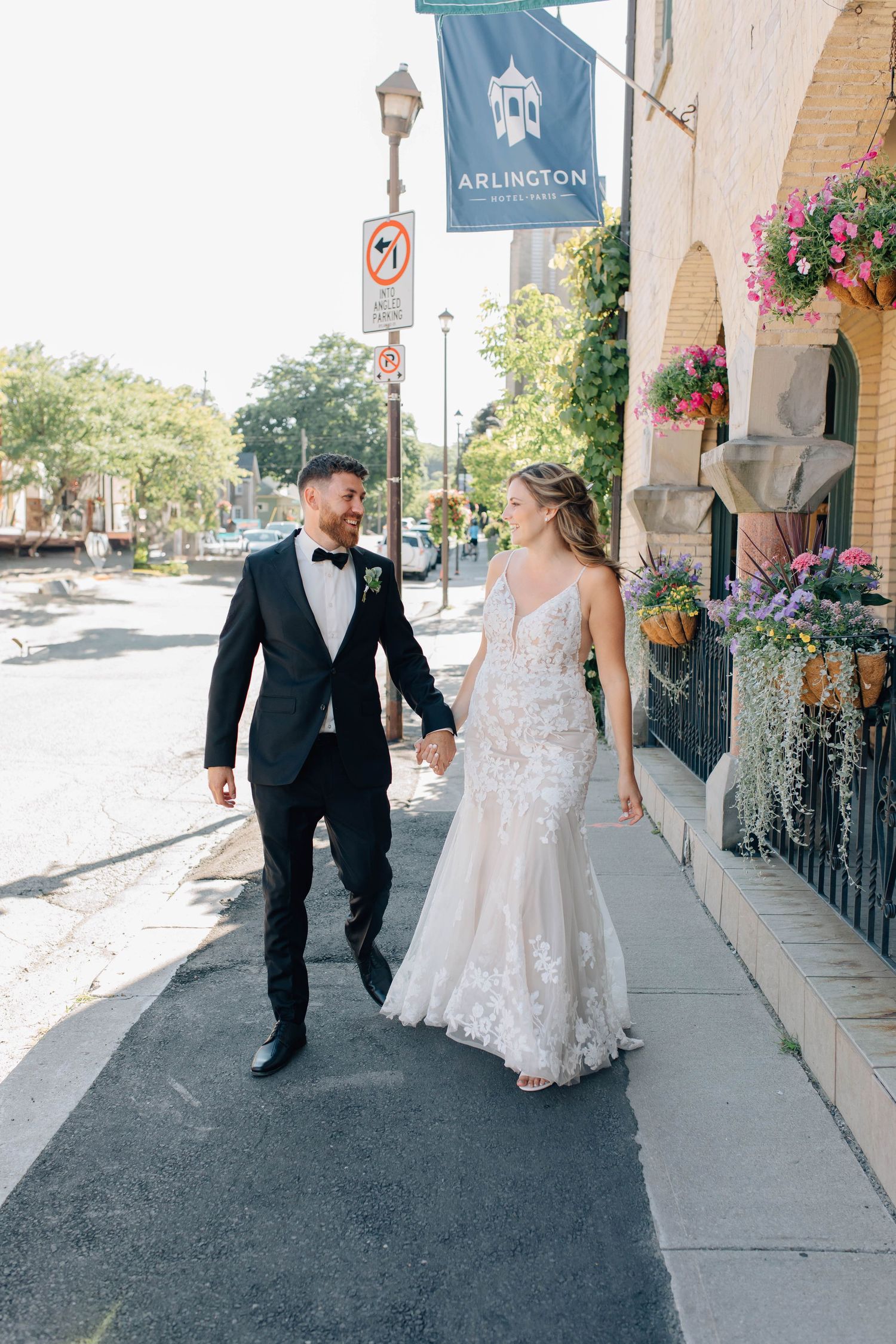 Bride and groom walk hand-in-hand on sunny Paris Ontario street, bride in fitted lace wedding gown.
