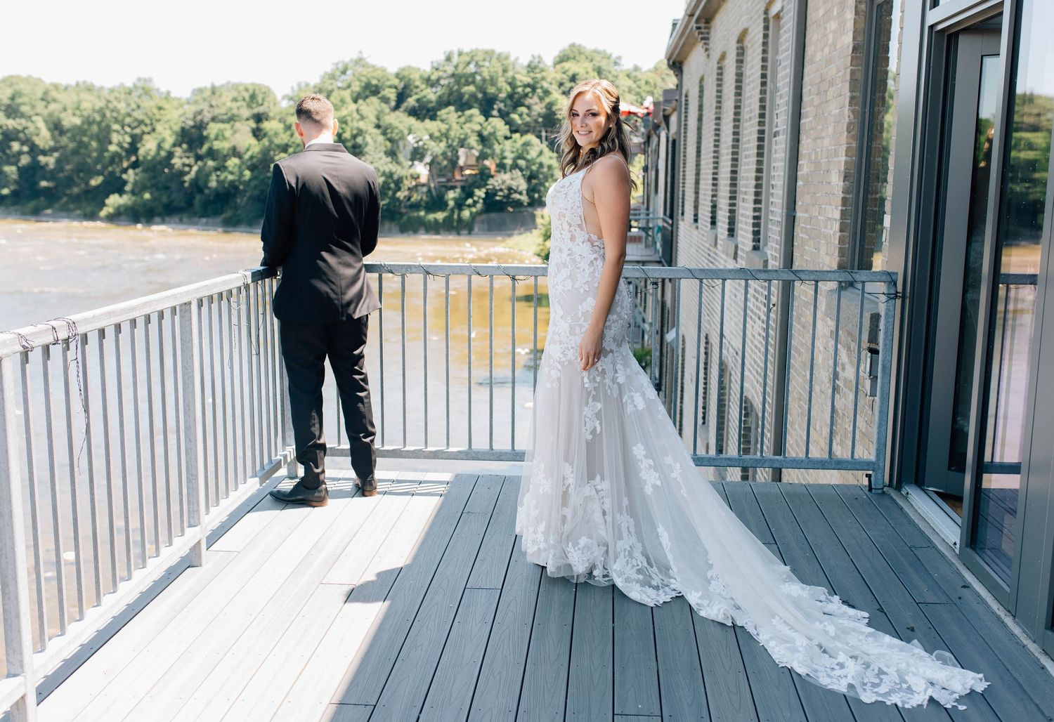 Bride in lace gown stands on balcony for first look reveal with groom outdoors.