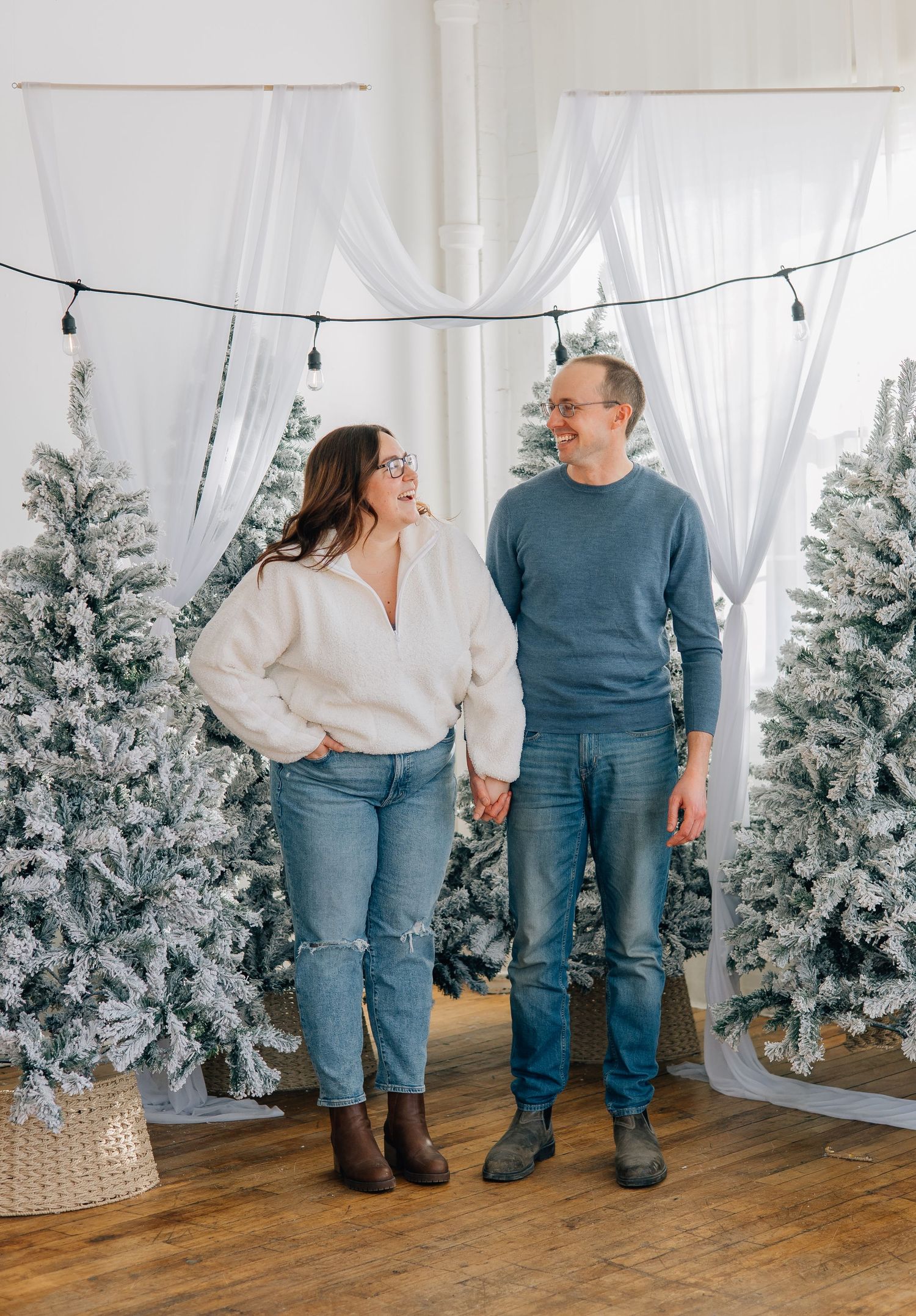 Couple holding hands laughing among snow-flocked Christmas trees in bright studio setting.