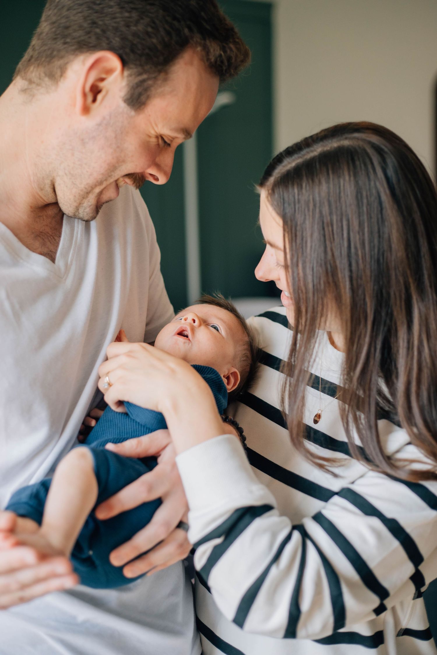New parents smiling lovingly at their newborn baby dressed in blue onesie held between them.