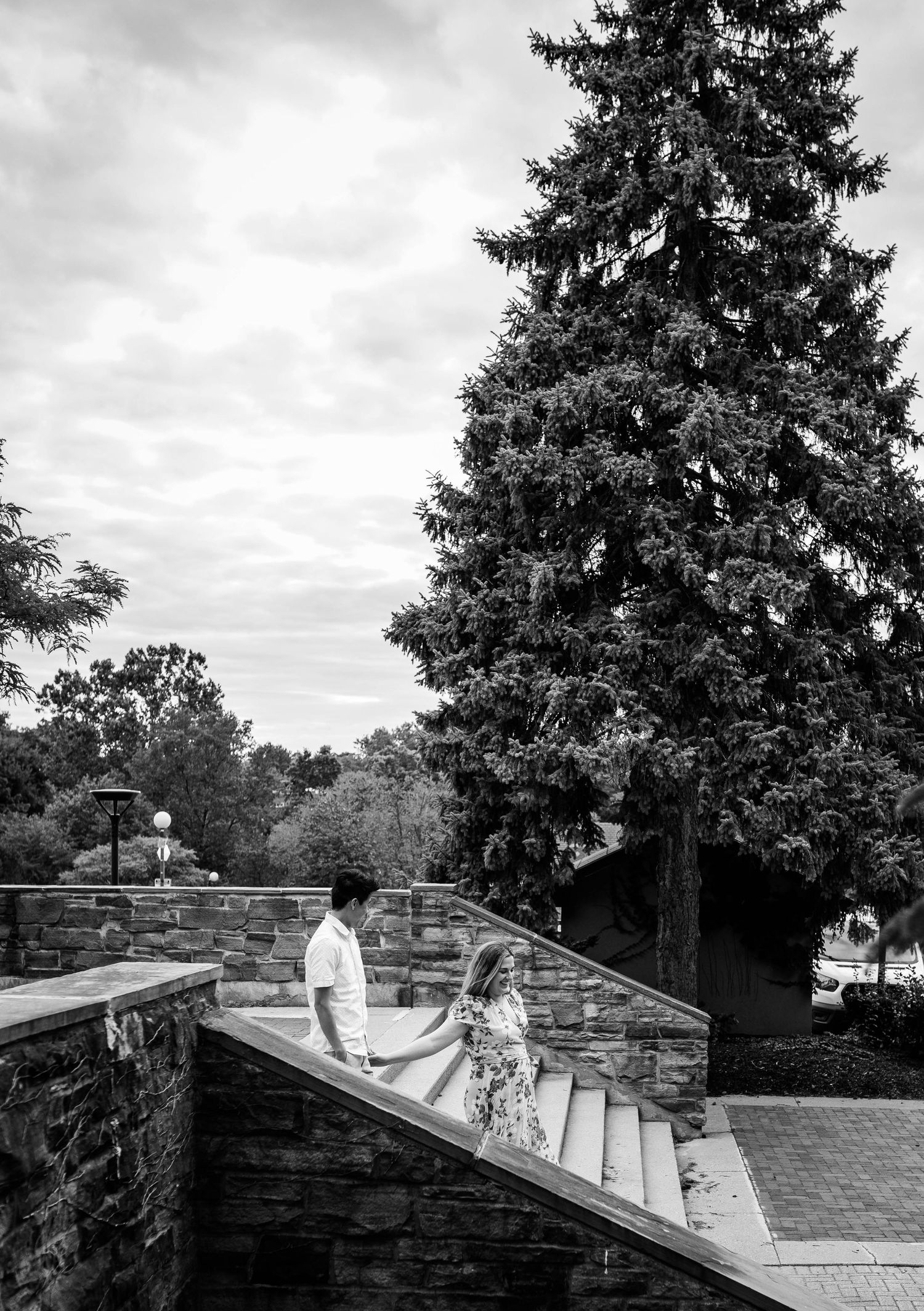 Black and white photo of couple holding hands on stone staircase with tall evergreen tree backdrop.