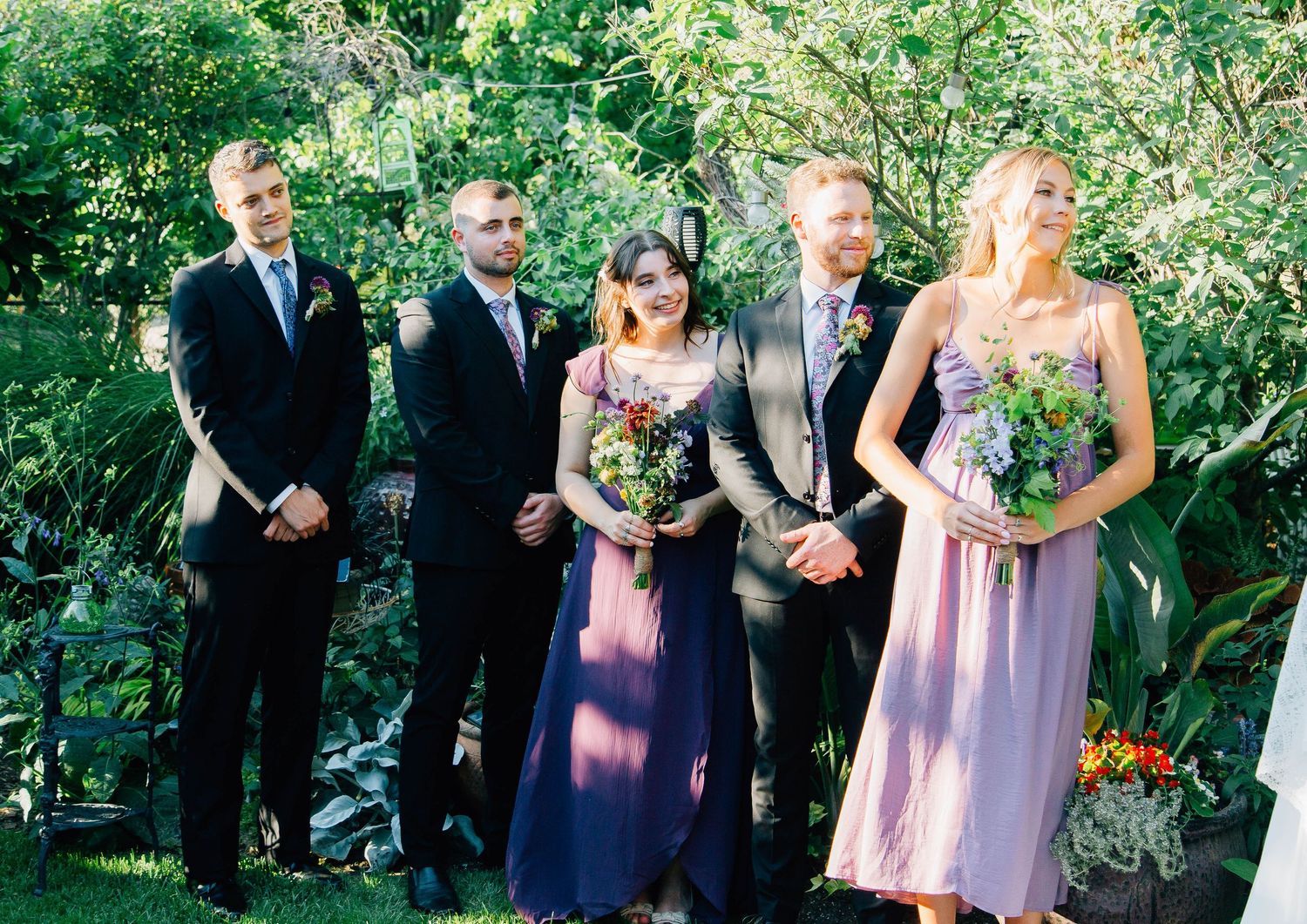 Wedding party stands at outdoor garden ceremony altar, groomsmen in suits and bridesmaids in purple and pink dresses.