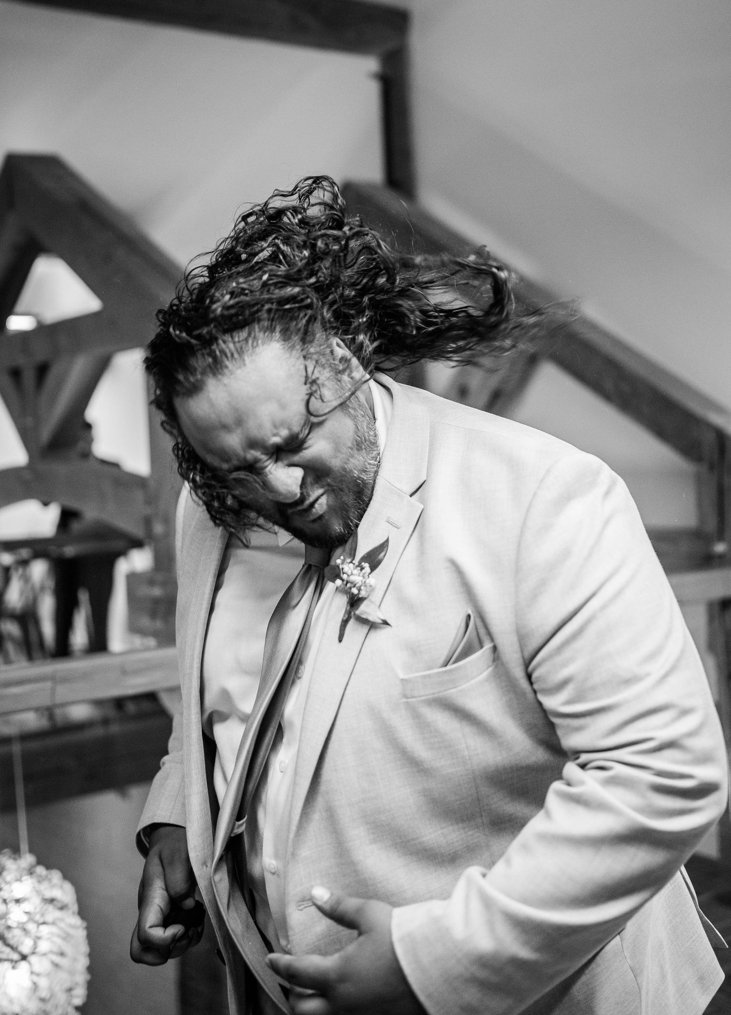 Black and white photo of groomsman in white suit laughing and bowing at wedding celebration.