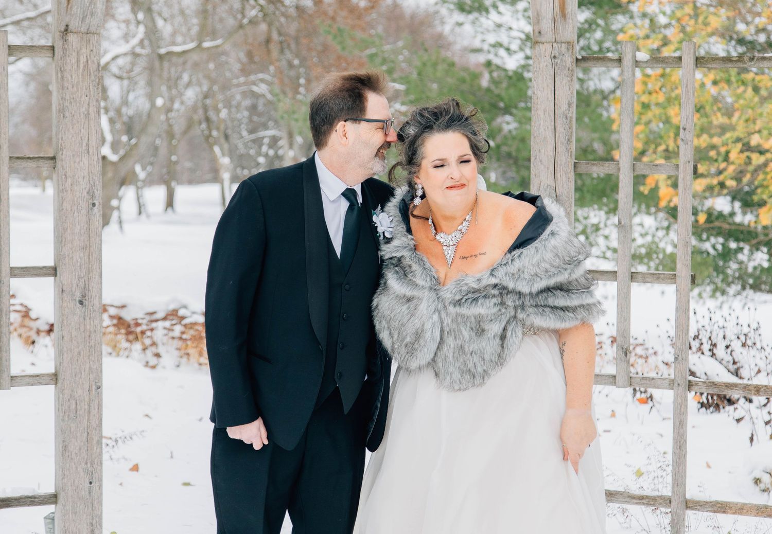 Bride in white gown with gray fur wrap and groom in black suit share a smile in winter snow.
