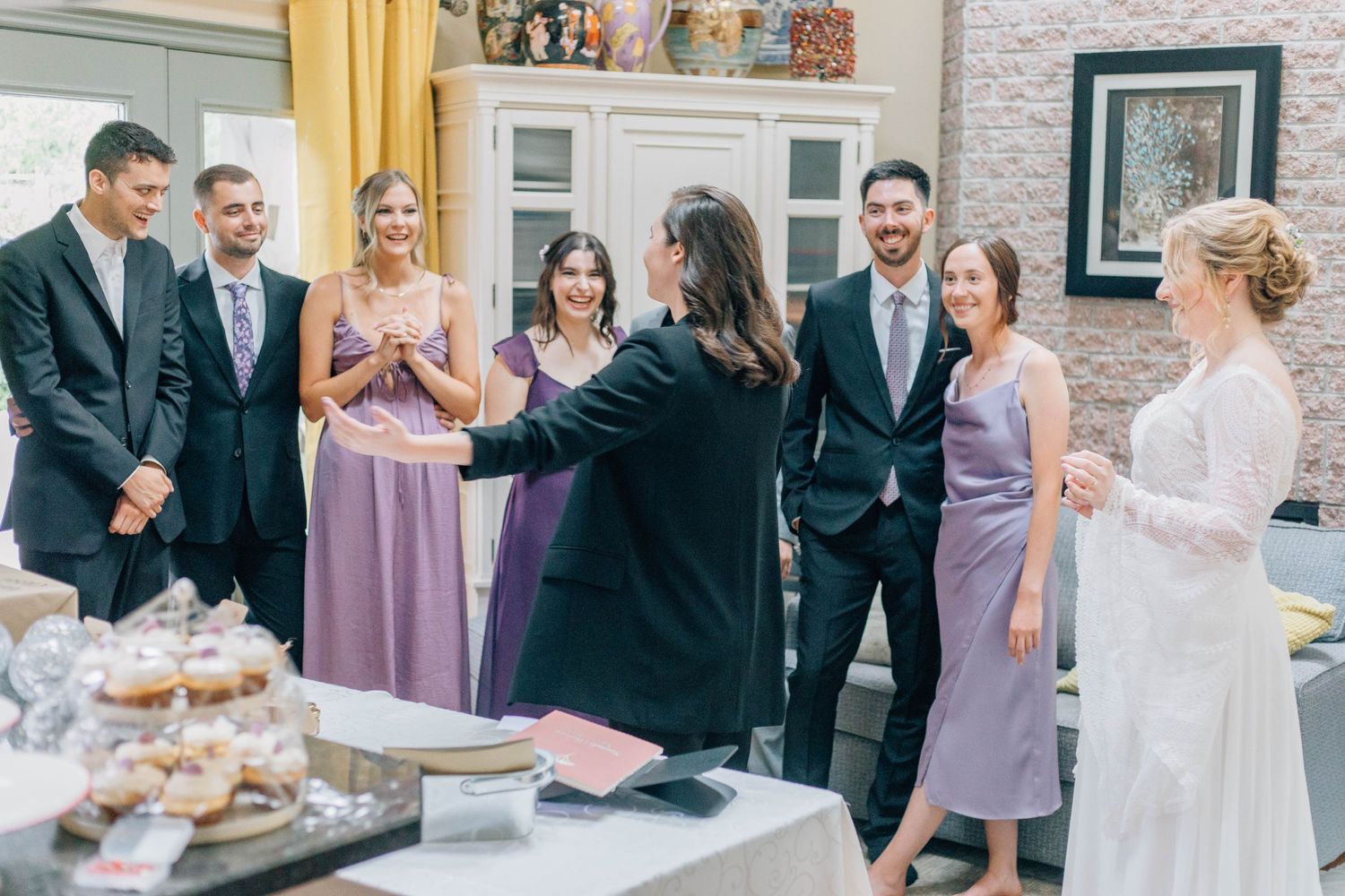 Bride and partner in black blazer greet smiling wedding guests in purple dresses indoors.