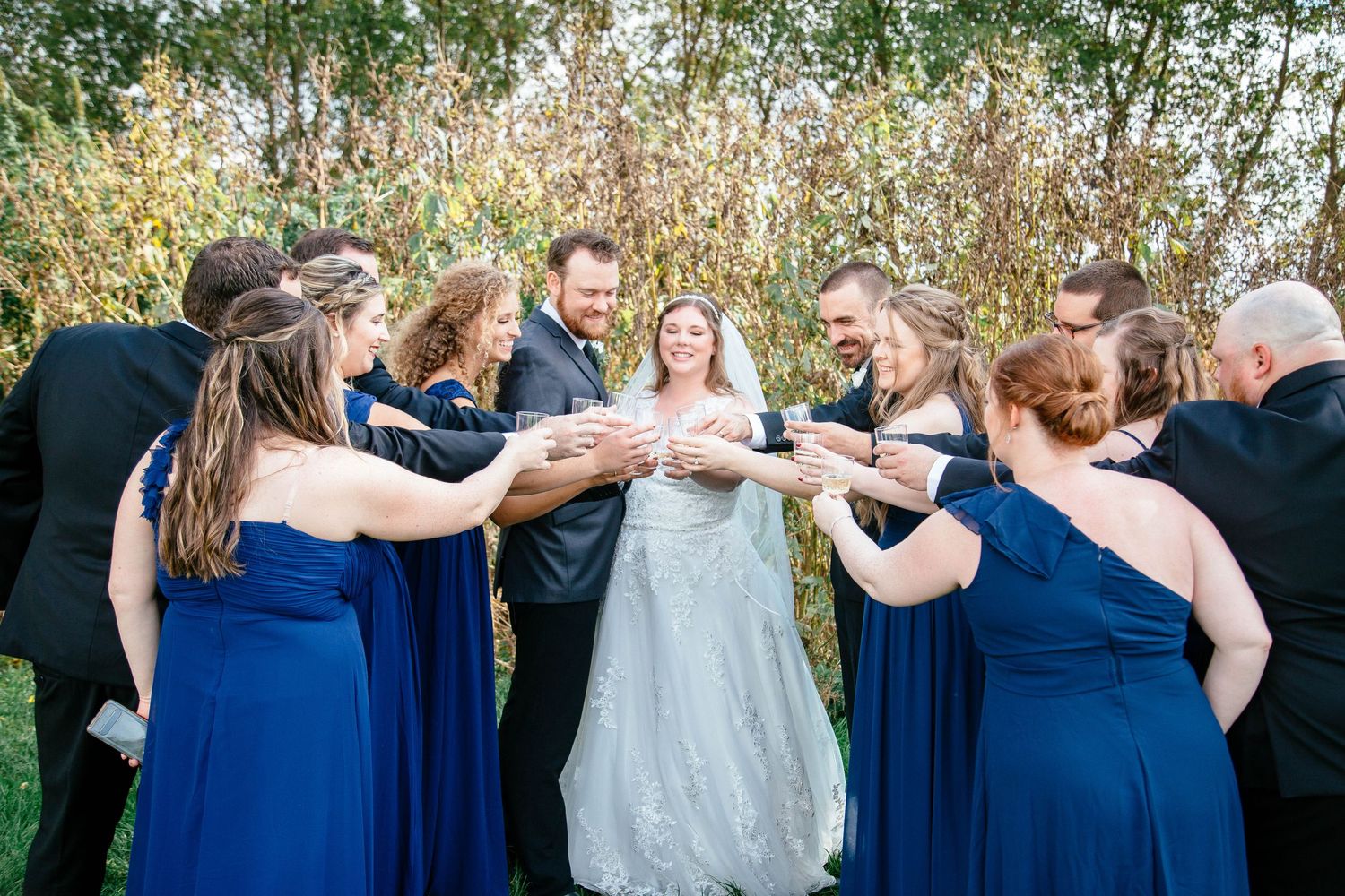 Bride and groom surrounded by wedding party in royal blue dresses toasting with champagne glasses.