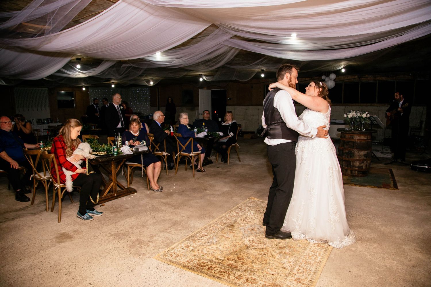 Bride and groom sharing first dance while wedding guests watch in a rustic barn reception hall.